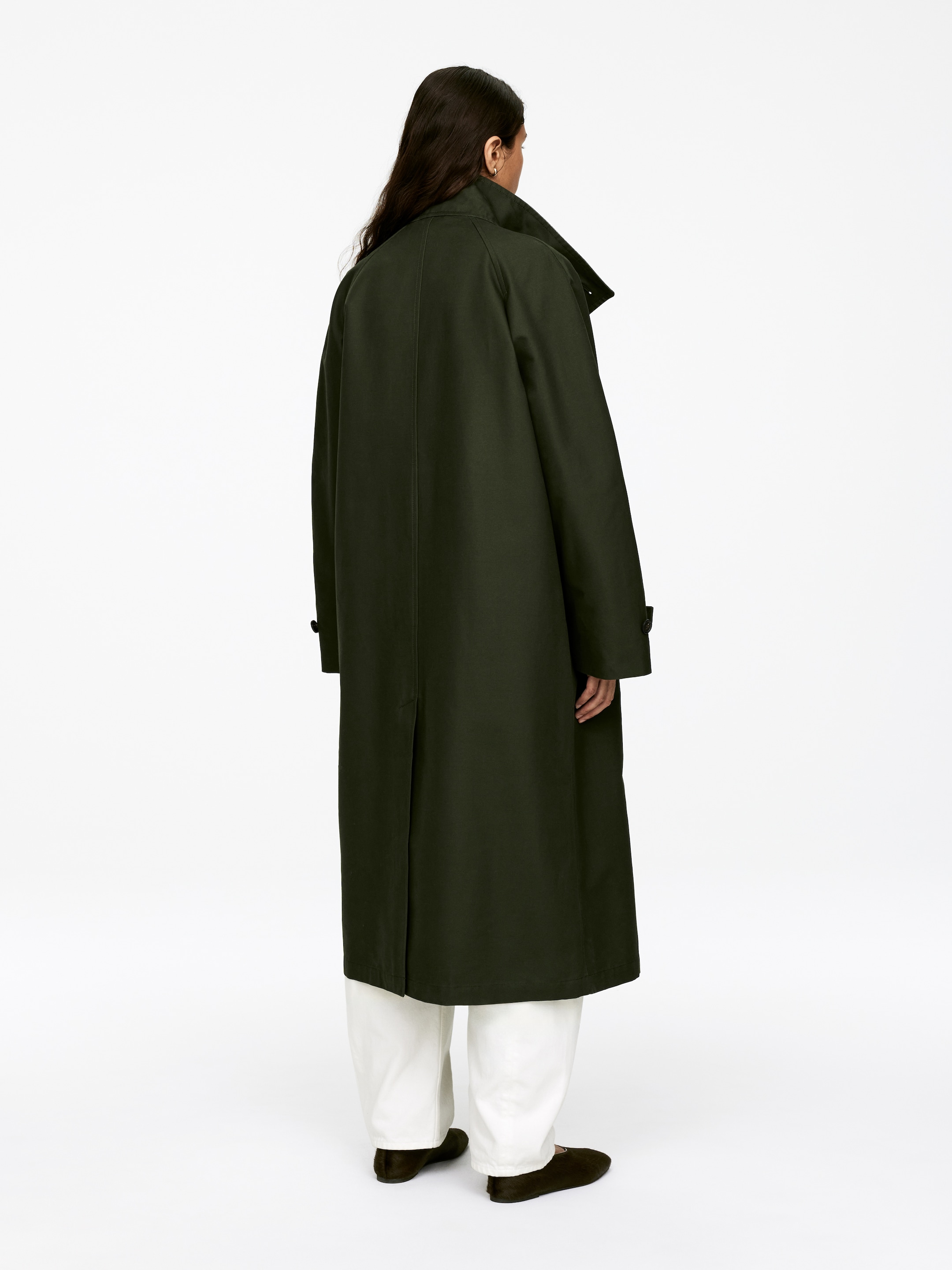 Visualizza immagine più grande: Un lungo cappotto verde scuro è presentato dalla vista posteriore, indossato sopra pantaloni bianchi. Presenta maniche raglan con linguette con bottoni sui polsini, un colletto rialzato e uno spacco centrale posteriore, che si estende significativamente oltre le ginocchia.