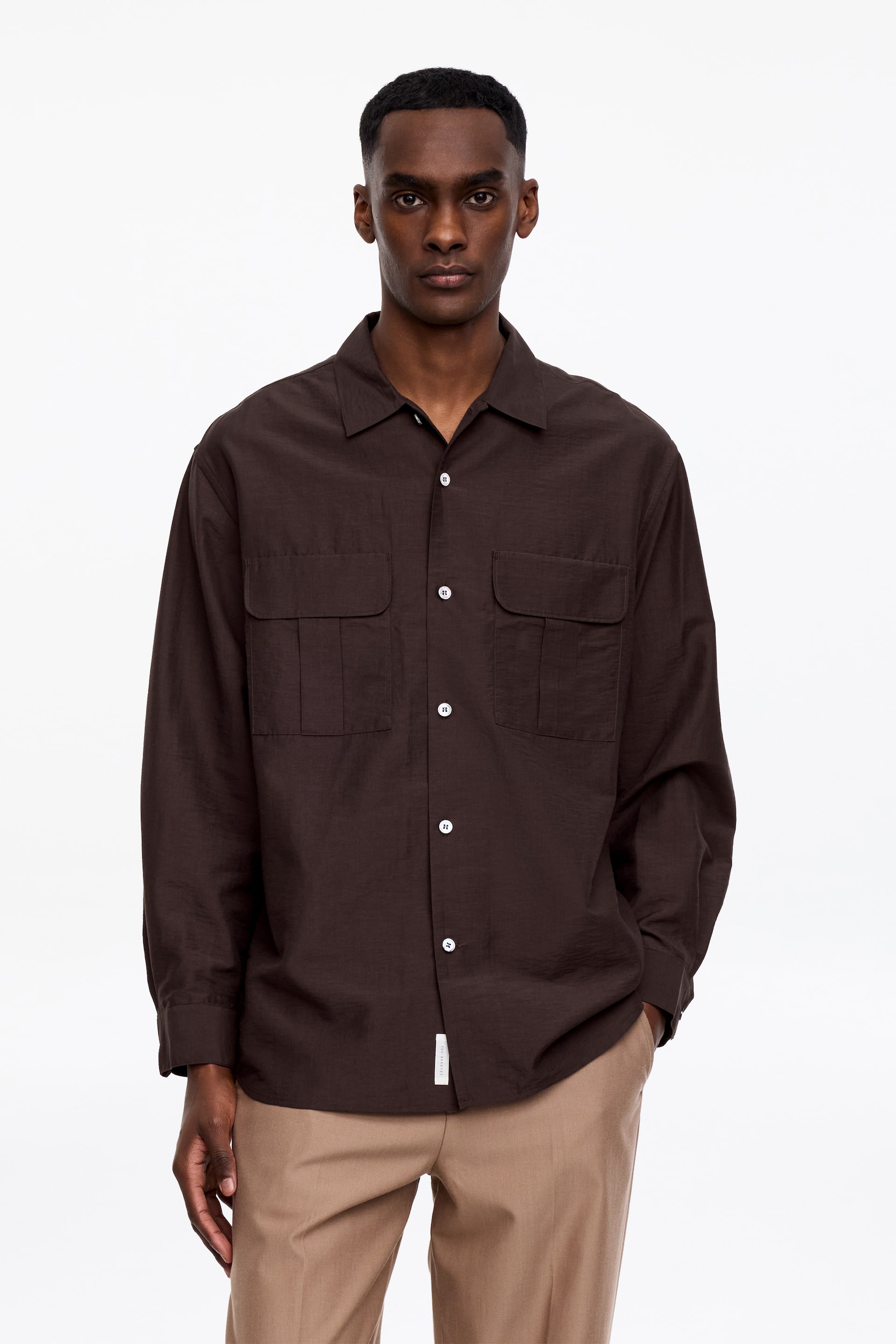 Visa större bild: En mörkbrun långärmad skjorta med en något skrynklig textur visas framifrån, med en spetsig krage, en fullknäppt framsida med vita knappar och två stora veckade påsydda fickor på bröstet. Skjortan har en avslappnad passform med långa ärmar, knäppta manschetter och en liten vit märkesetikett synlig på vänster fåll.