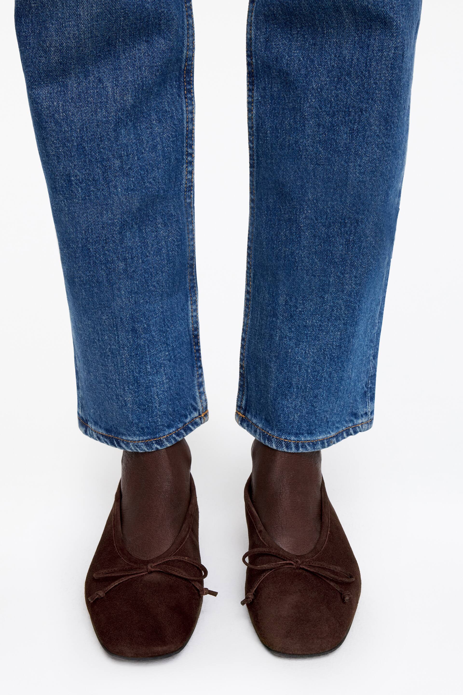 Προβολή μεγαλύτερης εικόνας: Ένα ζευγάρι σκούρα καφέ σουέντ μπαλαρίνες παρουσιάζεται, με μια μικρή λεπτομέρεια φιόγκου στο επάνω μέρος κάθε παπουτσιού. Τα παπούτσια είναι στραμμένα προς τα εμπρός, προβάλλοντας κάτω από το στρίφωμα ενός μπλε τζιν.