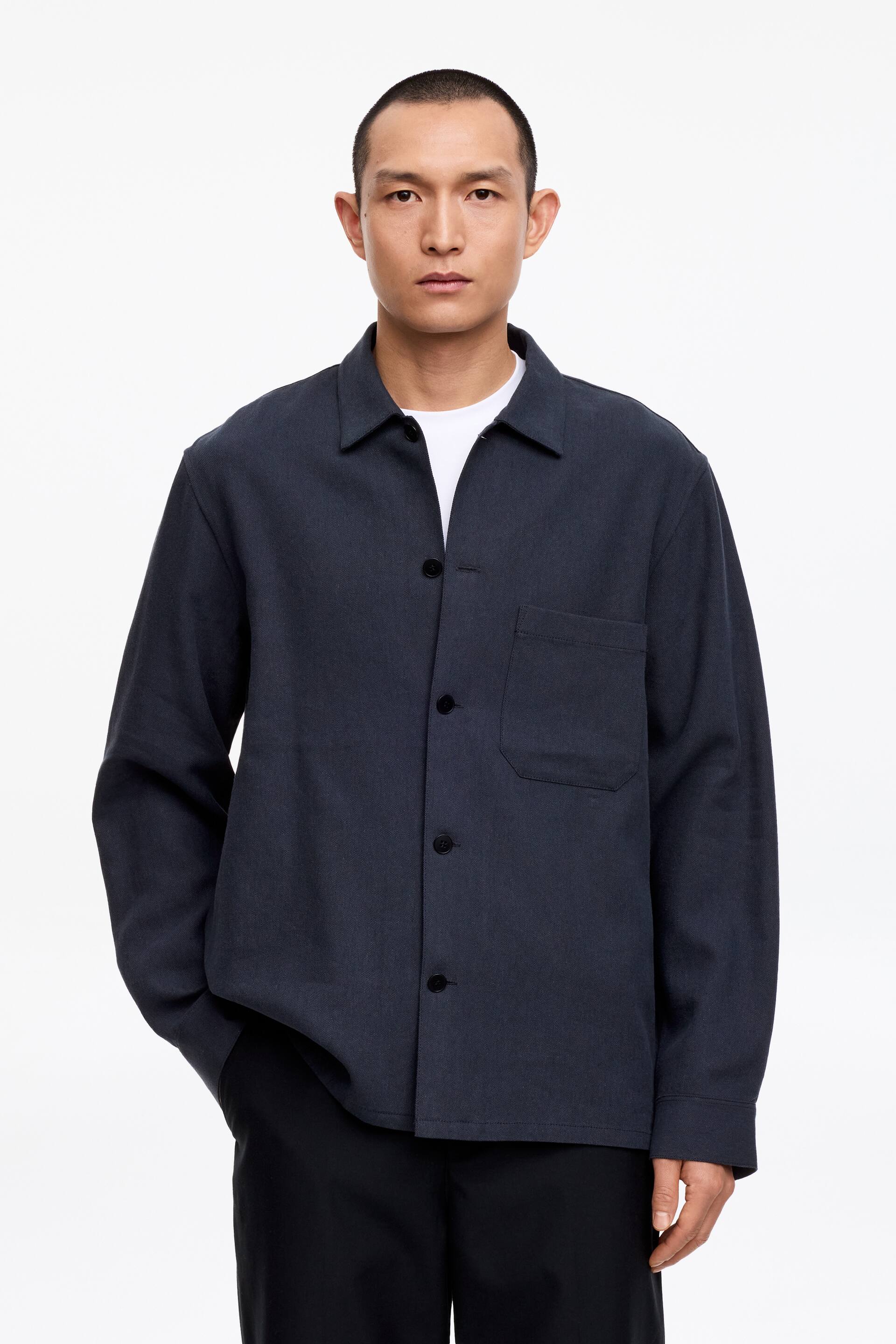 Agrandir l'image: La chemise texturée gris foncé présente un col à pointes écartées, une fermeture boutonnée sur le devant avec des boutons foncés et une poche plaquée simple sur le côté gauche de la poitrine. Elle est portée par-dessus un t-shirt blanc.