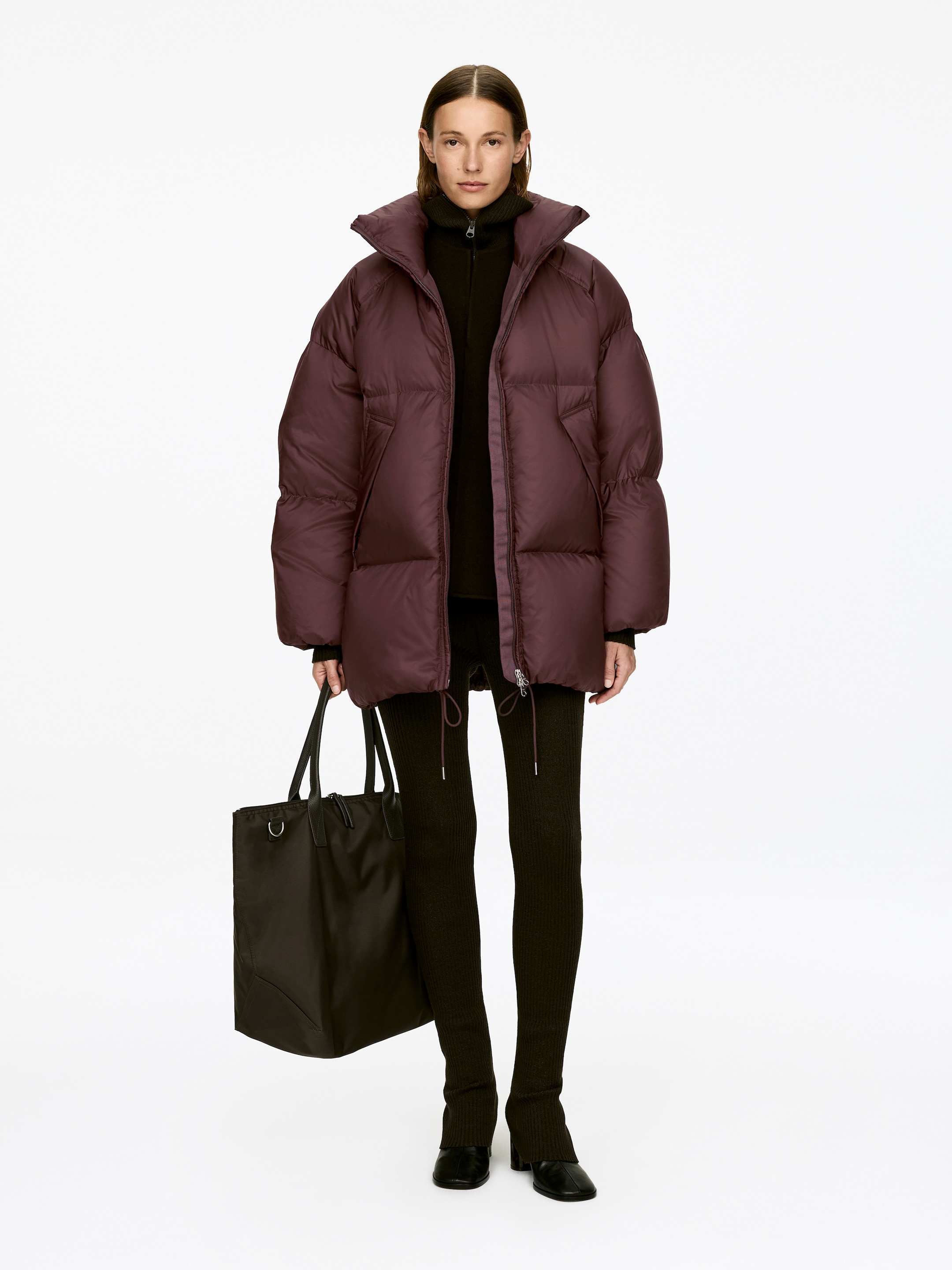 View larger image: Upcycled Down Puffer Jacket, a dark plum, oversized design with horizontal quilting and a high stand collar, is being worn by the model. It is layered over a dark high-neck top, and the model is carrying a dark tote bag.