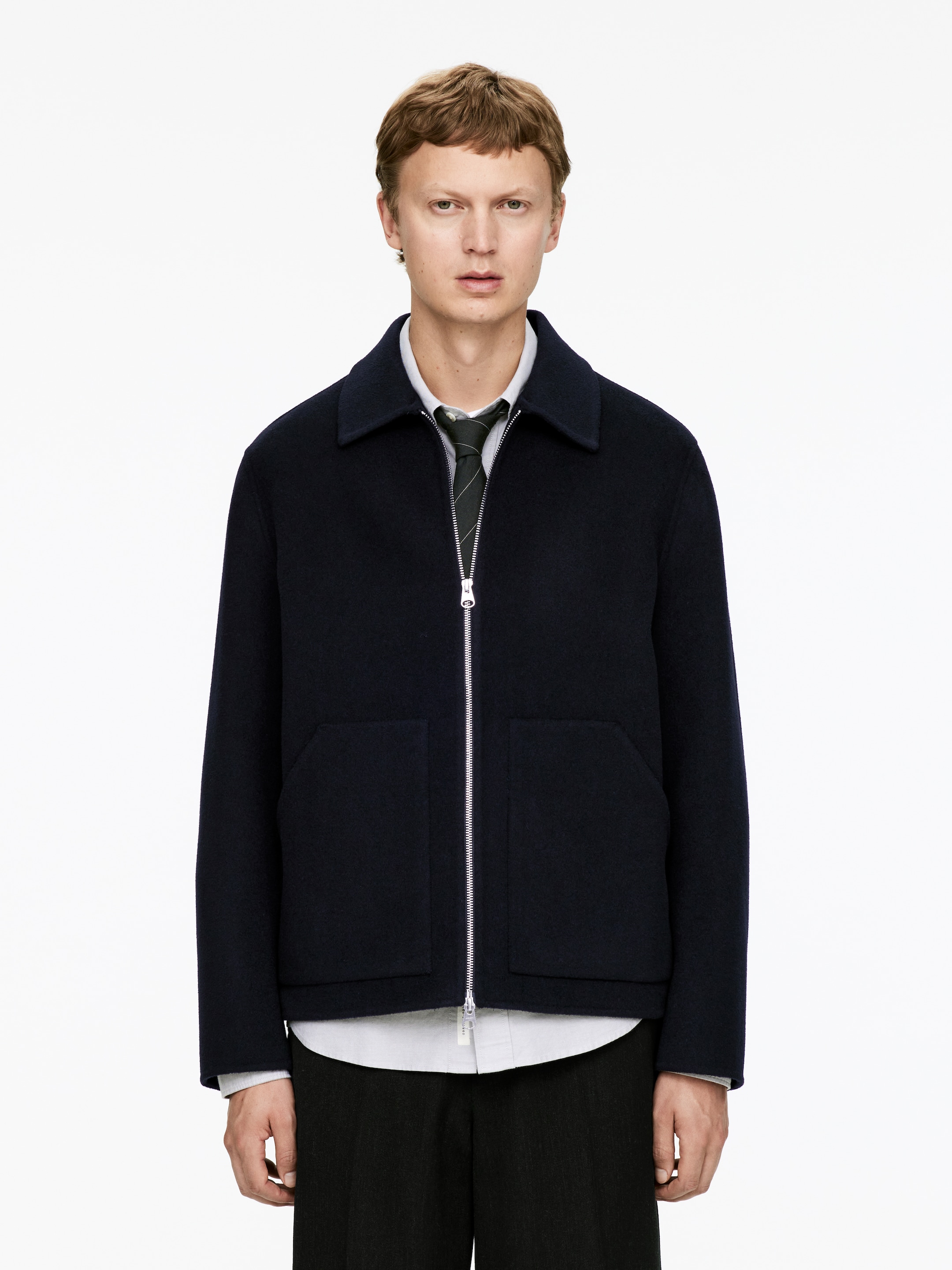 Grösseres Bild ansehen: Eine marineblaue Blousonjacke wird von vorne präsentiert, mit einem spitzen Kragen, einem silberfarbenen Reißverschluss und zwei großen rechteckigen aufgesetzten Taschen. Sie wird über einem hellen Hemd mit Kragen und einer dunklen gestreiften Krawatte getragen.