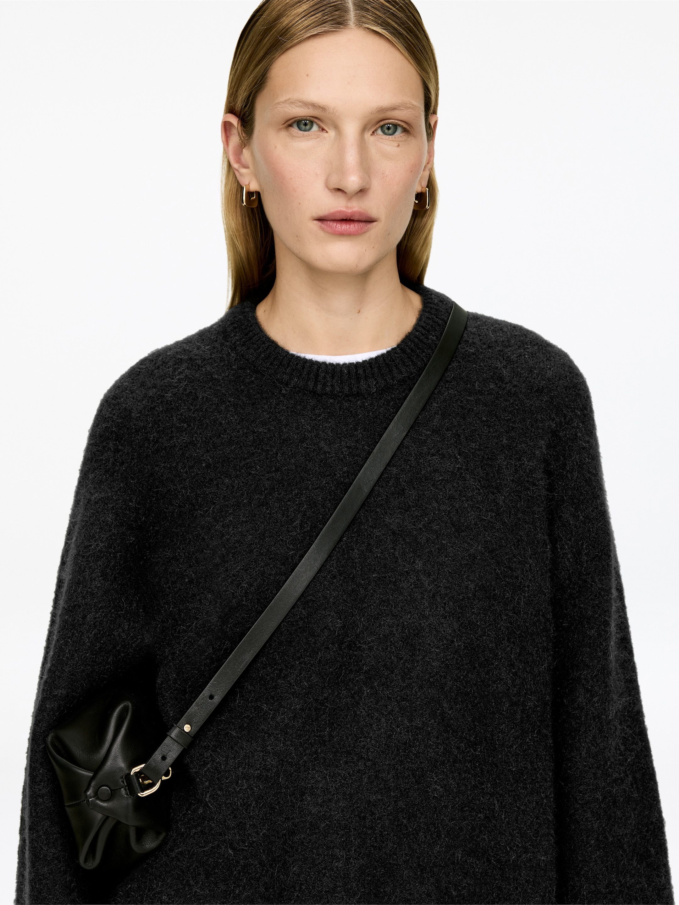 Grösseres Bild ansehen: Ein anthrazitgrauer, flauschiger Strickpullover mit Rundhalsausschnitt wird von vorne getragen, zeichnet sich durch eine entspannte Passform und lange Ärmel aus. Ein weißes T-Shirt mit Rundhalsausschnitt ist am Kragen sichtbar, und eine kleine schwarze Ledertasche mit dünnem Gurt trägt über dem Pullover.