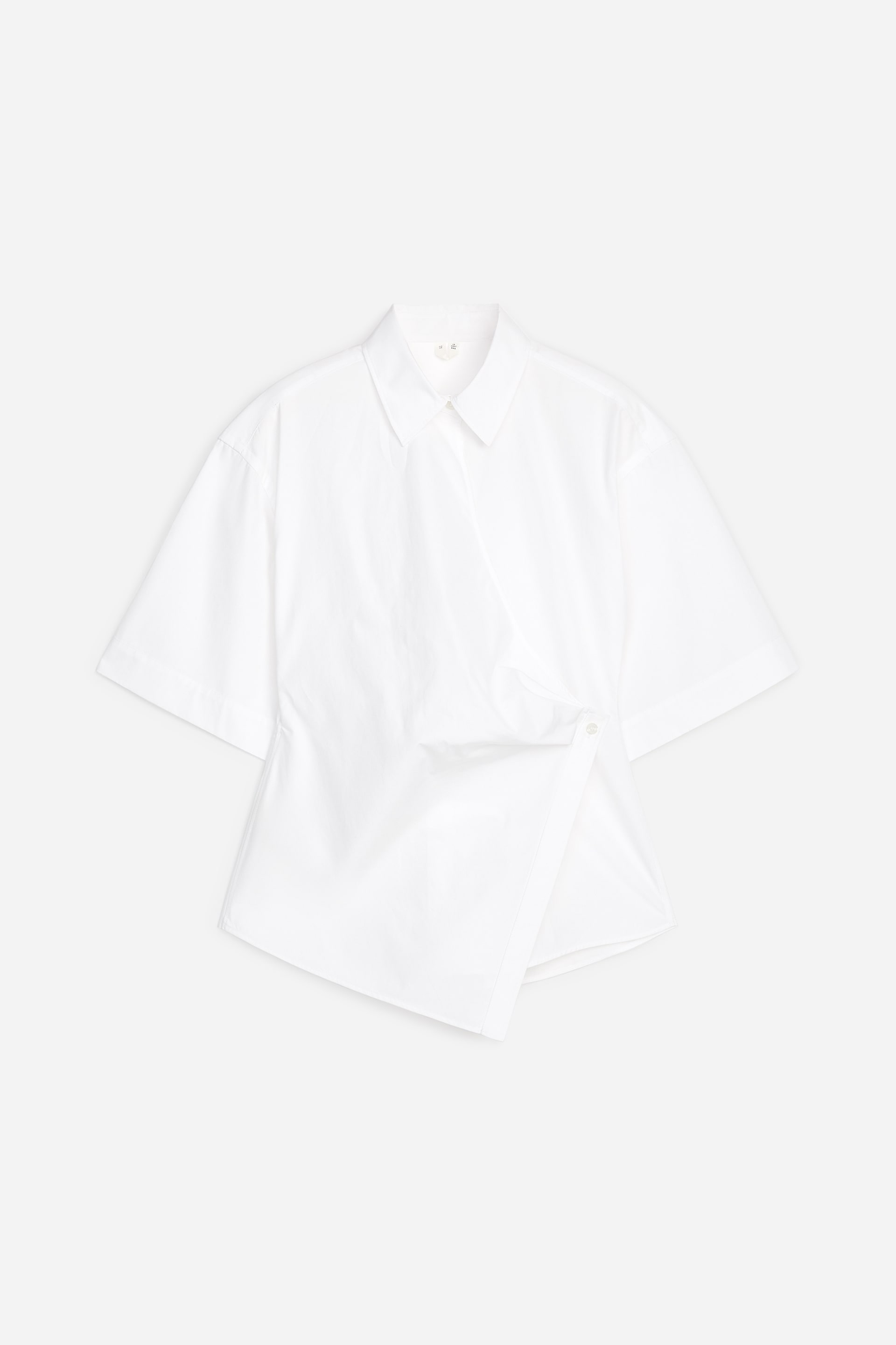 Ver imagen más grande: Una camisa blanca con cuello en punta, mangas cortas y un diseño de envoltura frontal. La envoltura se asegura con un único botón blanco en el lado derecho, creando un dobladillo asimétrico.