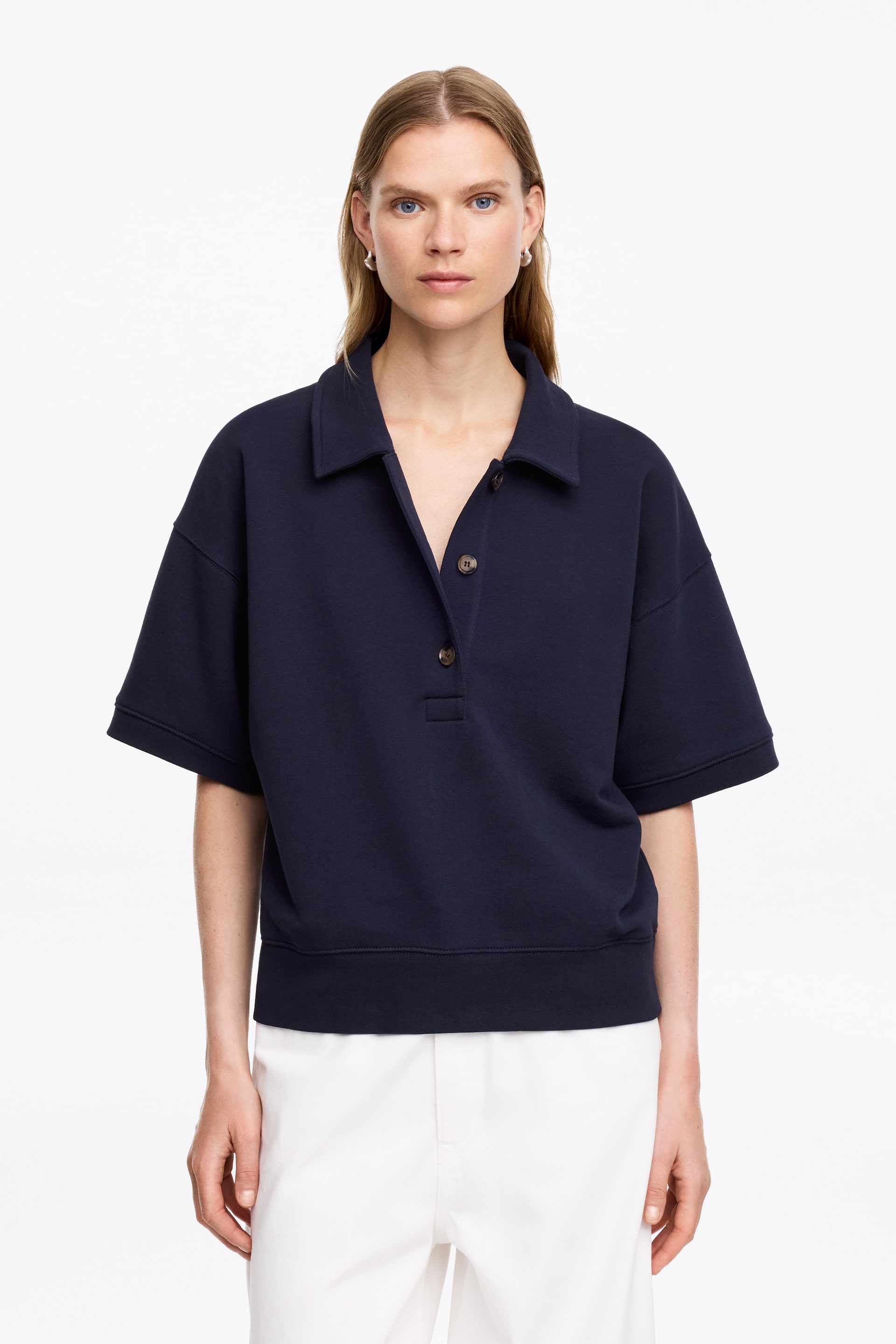 Agrandir l'image: Un sweat-shirt de style polo bleu marine est présenté de face, avec un col étalé et une patte de boutonnage à trois boutons marron foncé, dont deux sont déboutonnés. Il arbore des manches courtes et larges ainsi qu'une bande d'ourlet côtelée.