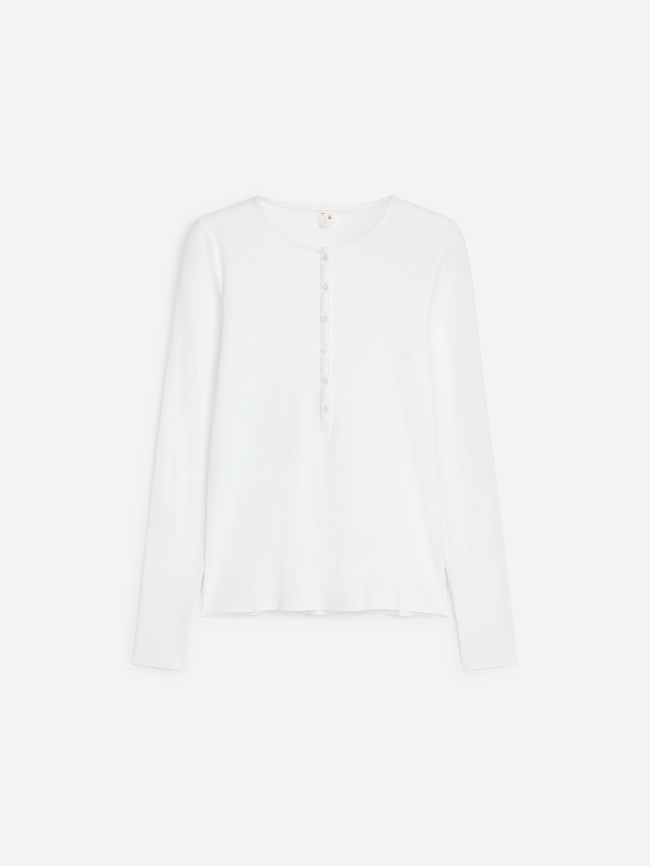 Das Model trÃ¤gt ein weiÃes Henley-Top mit langen Ãrmeln und Rippstruktur, mit schmaler Knopfleiste und kleinen KnÃ¶pfen, die am Kragen teilweise offen gelassen sind. Das Top ist in eine schwarze, taillierte Hose gesteckt.