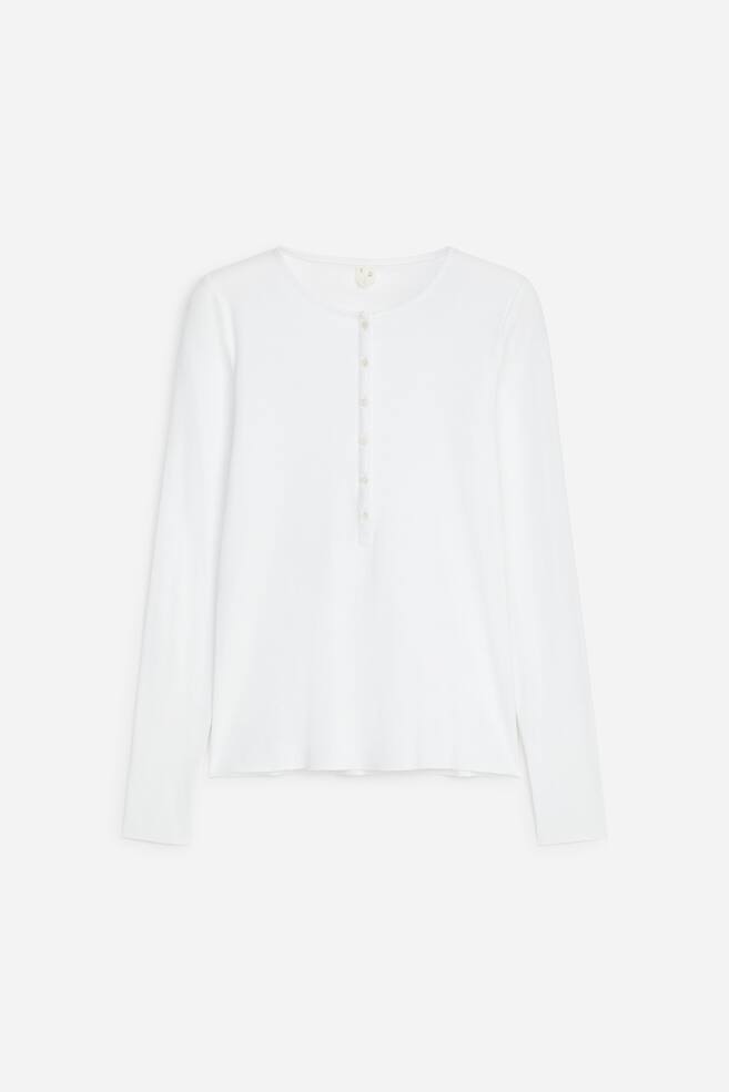 Ein weiÃes Henley-Top mit langen Ãrmeln ist von vorne zu sehen und verfÃ¼gt Ã¼ber einen runden Ausschnitt sowie eine vertikale Knopfleiste mit sechs kleinen KnÃ¶pfen. Das Top zeigt lange, schmale Ãrmel und einen geraden Saum mit feiner Strickstruktur. - 1