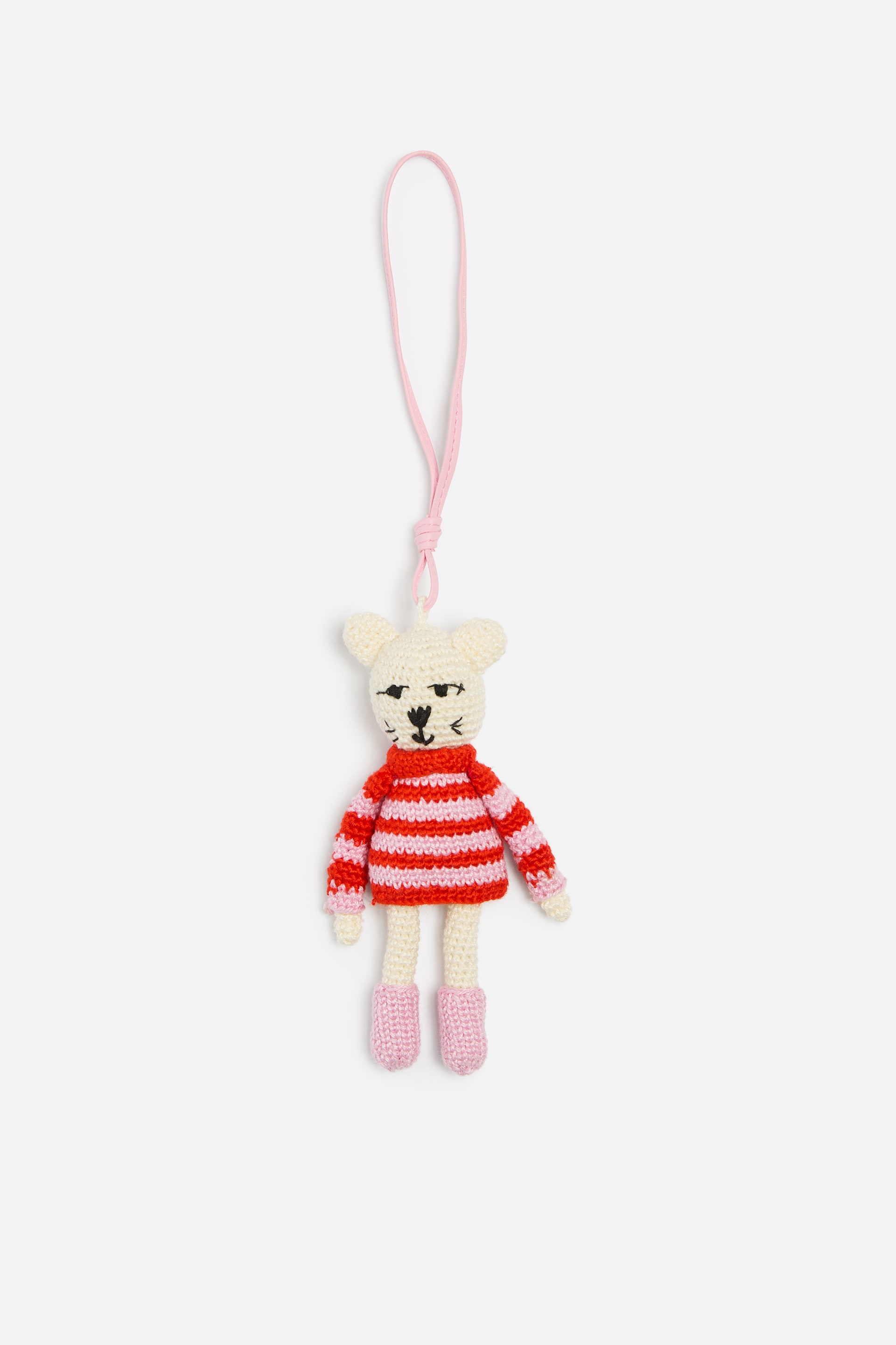 Ver imagen más grande: Un colgante de bolso de animal tejido a crochet cuelga en posición vertical, de frente, y presenta un cuerpo de color crema con rasgos faciales negros bordados. La figura lleva un jersey a rayas rojas y rosas con botas rosas a juego, y está suspendida por una larga y fina trabilla rosa unida a la cabeza.