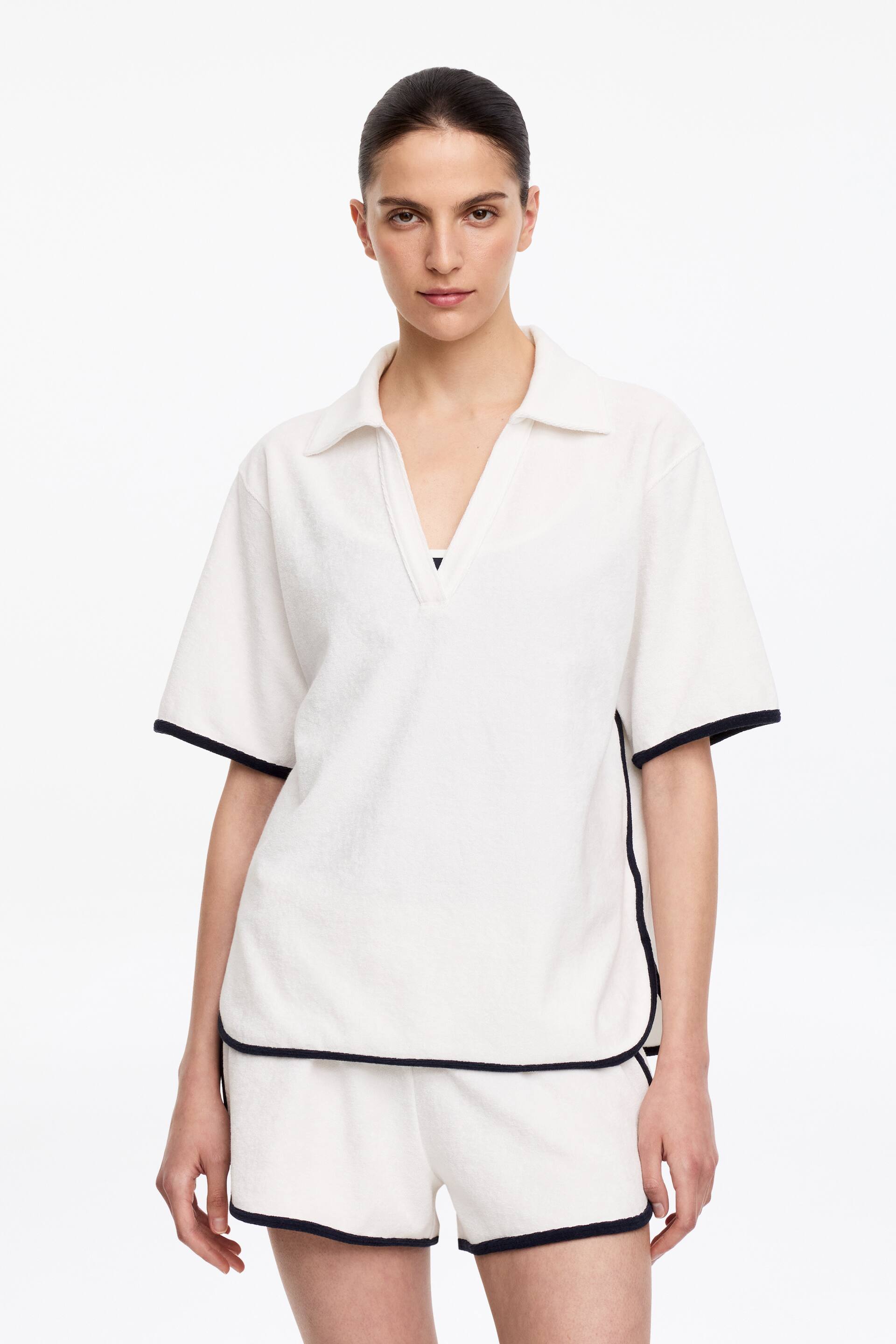 Ver imagen más grande: Se presenta un conjunto de dos piezas de tela de toalla blanca, orientado hacia adelante. Incluye una camiseta tipo polo de manga corta con cuello extendido y escote en V, y pantalones cortos a juego, ambos detallados con ribetes negros a lo largo de los bordes y costuras laterales.