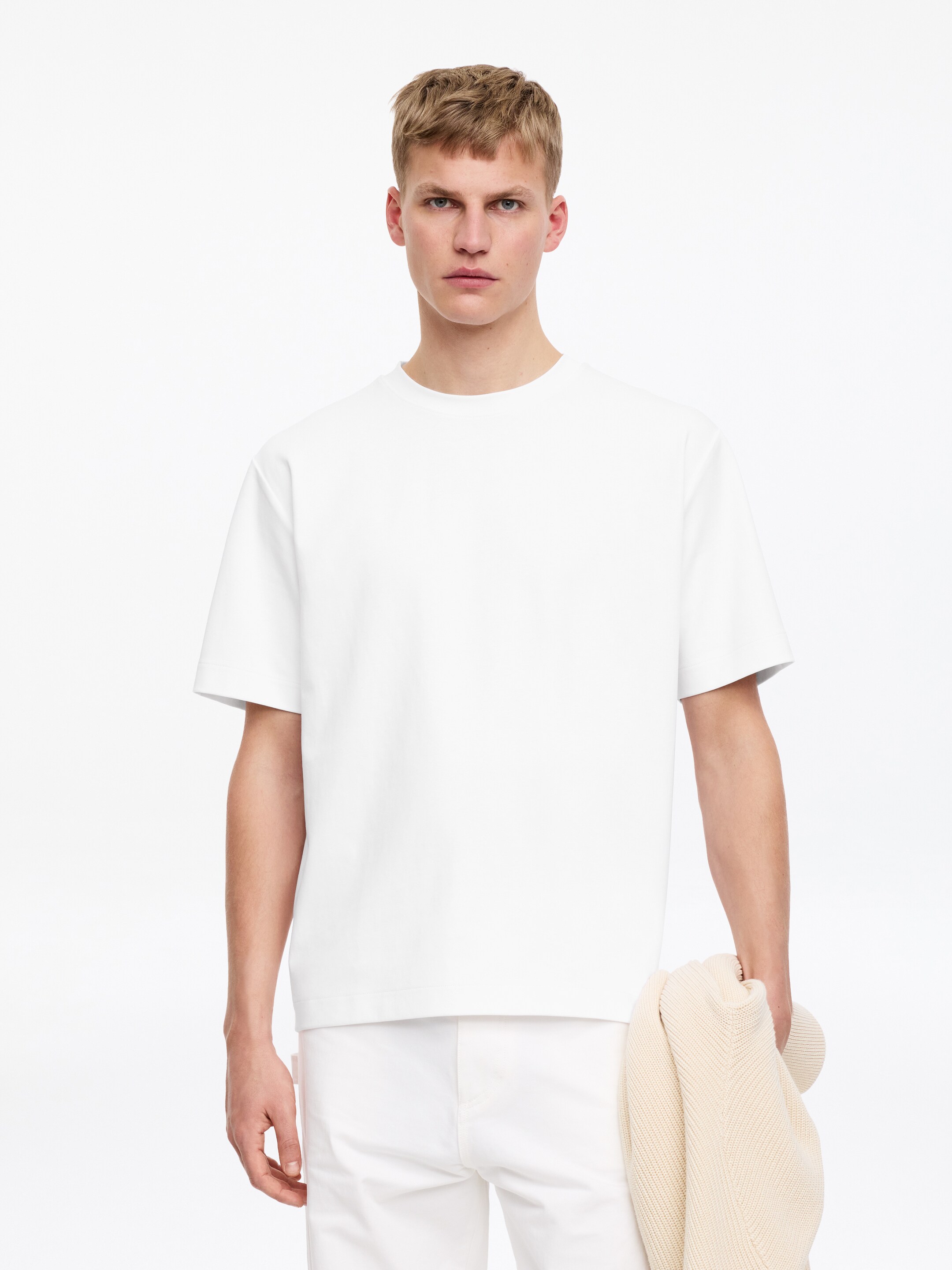 Visa större bild: Modellen bär en vit T-shirt med crewneck, regular fit och korta ärmar. Modellen bär även vita byxor och håller ett ljusfärgat stickat plagg i ena handen.