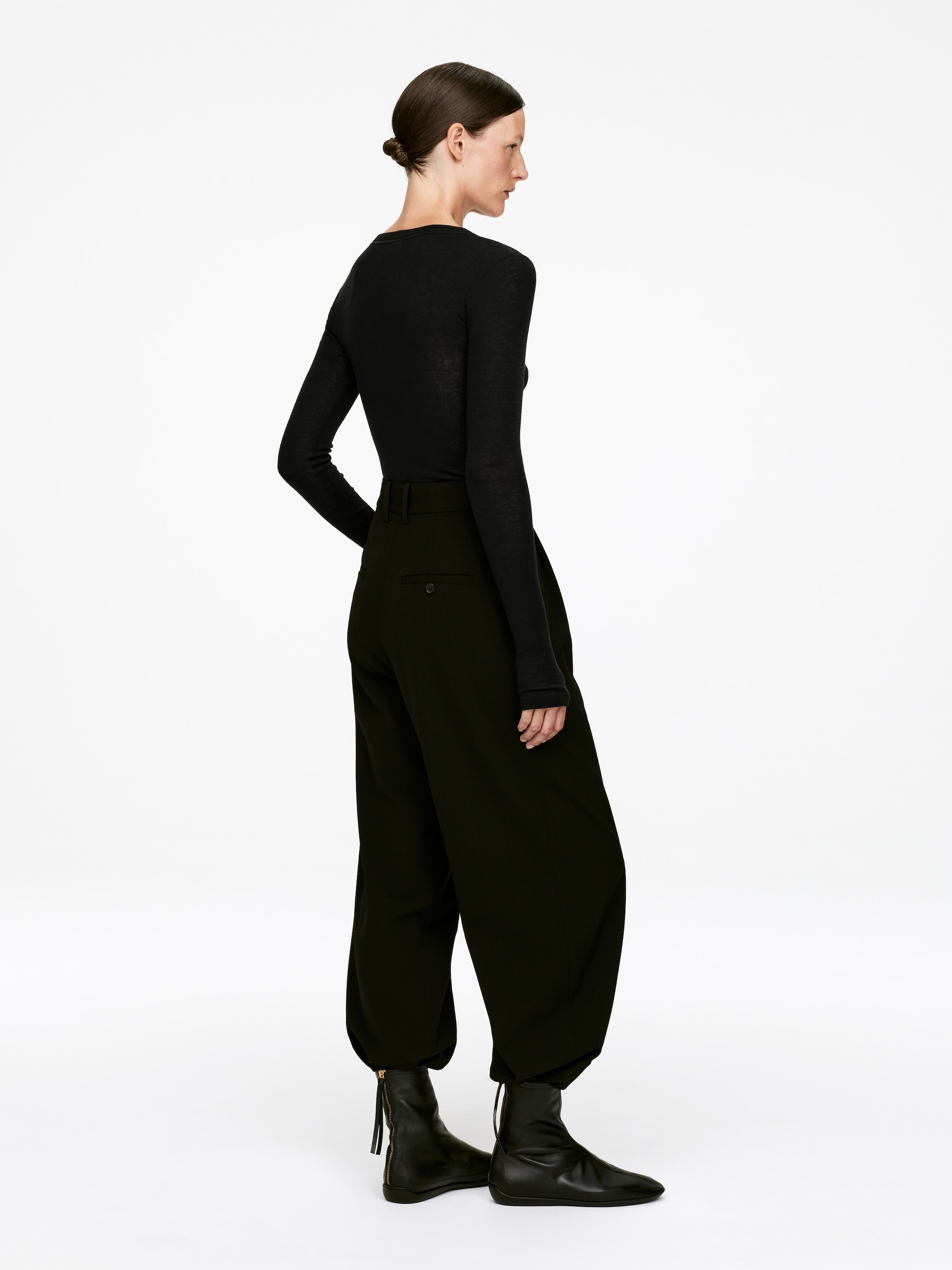 Se større billede: En sort langærmet, slim-fit top og sorte højtaljede, brede bukser bliver modelleret fra en sidevinkel. Bukserne samler sig ved anklen over sorte ankelstøvler, som har en synlig messingfarvet side lynlås.