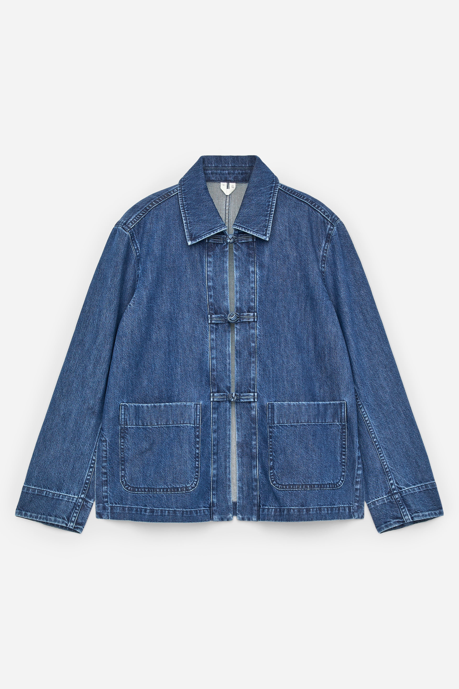 Grotere afbeelding bekijken: Een middelblauw denim jack is van voren te zien, met een spreidkraag en twee donkere kikker sluitingen aan de voorkant. Het heeft twee grote rechthoekige opgestikte zakken bij de taille en lange mouwen.