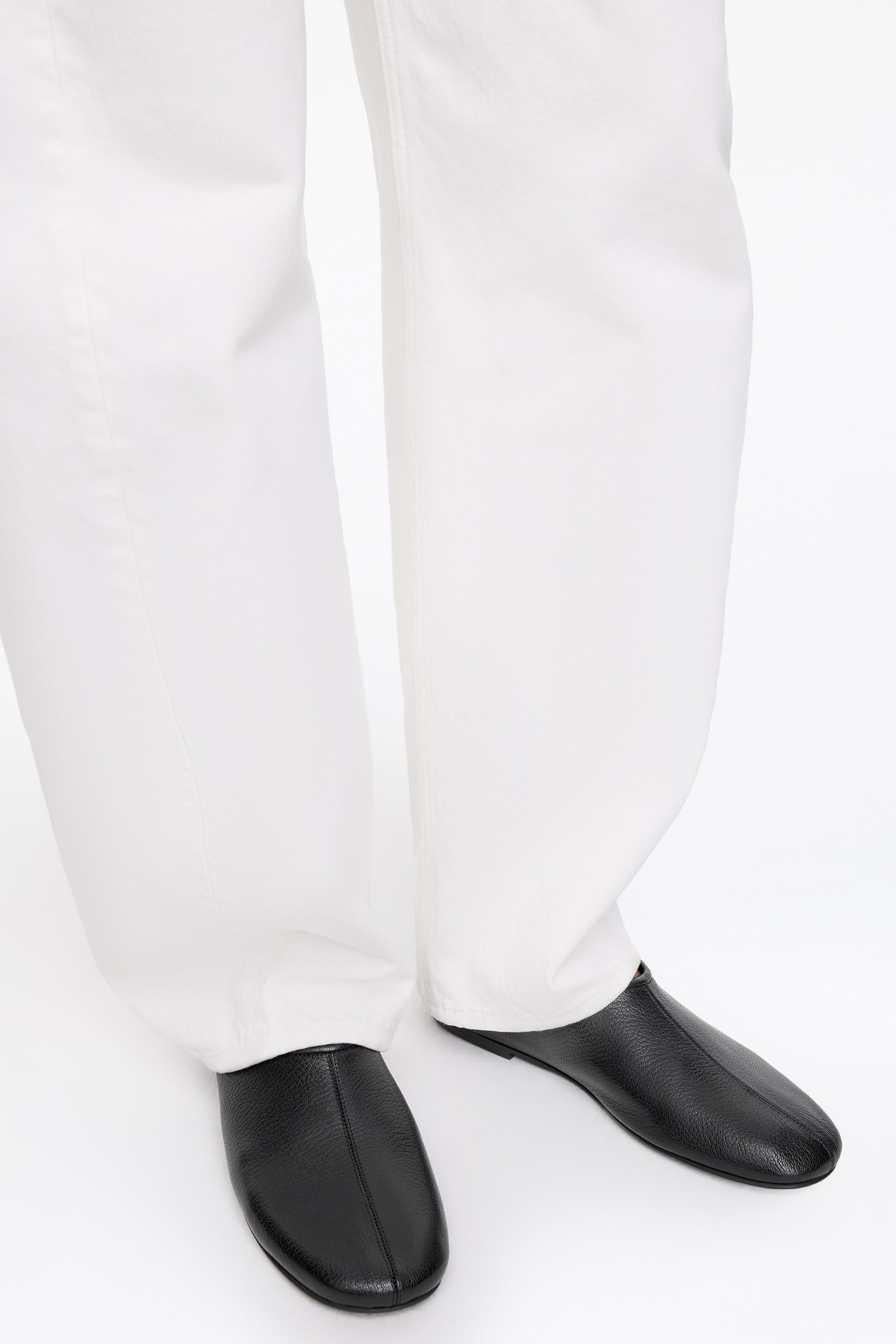 Visa större bild: Svarta läderslippers med en texturerad yta och en central söm på tån, presenterade från en något upphöjd frontvy. De bärs tillsammans med vita vida byxor som faller lätt över vristen.
