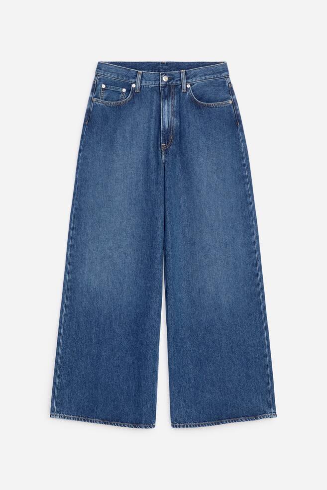 CEDAR Oversized Wide Jeans worden gepresenteerd in een vooraanzicht en hebben een medium blauwe wassing, een hoge taille en een wijde pijp. Ze tonen een voorste knoop- en ritssluiting, riemlussen en klassieke voorzakken met metalen klinknagels. - 1