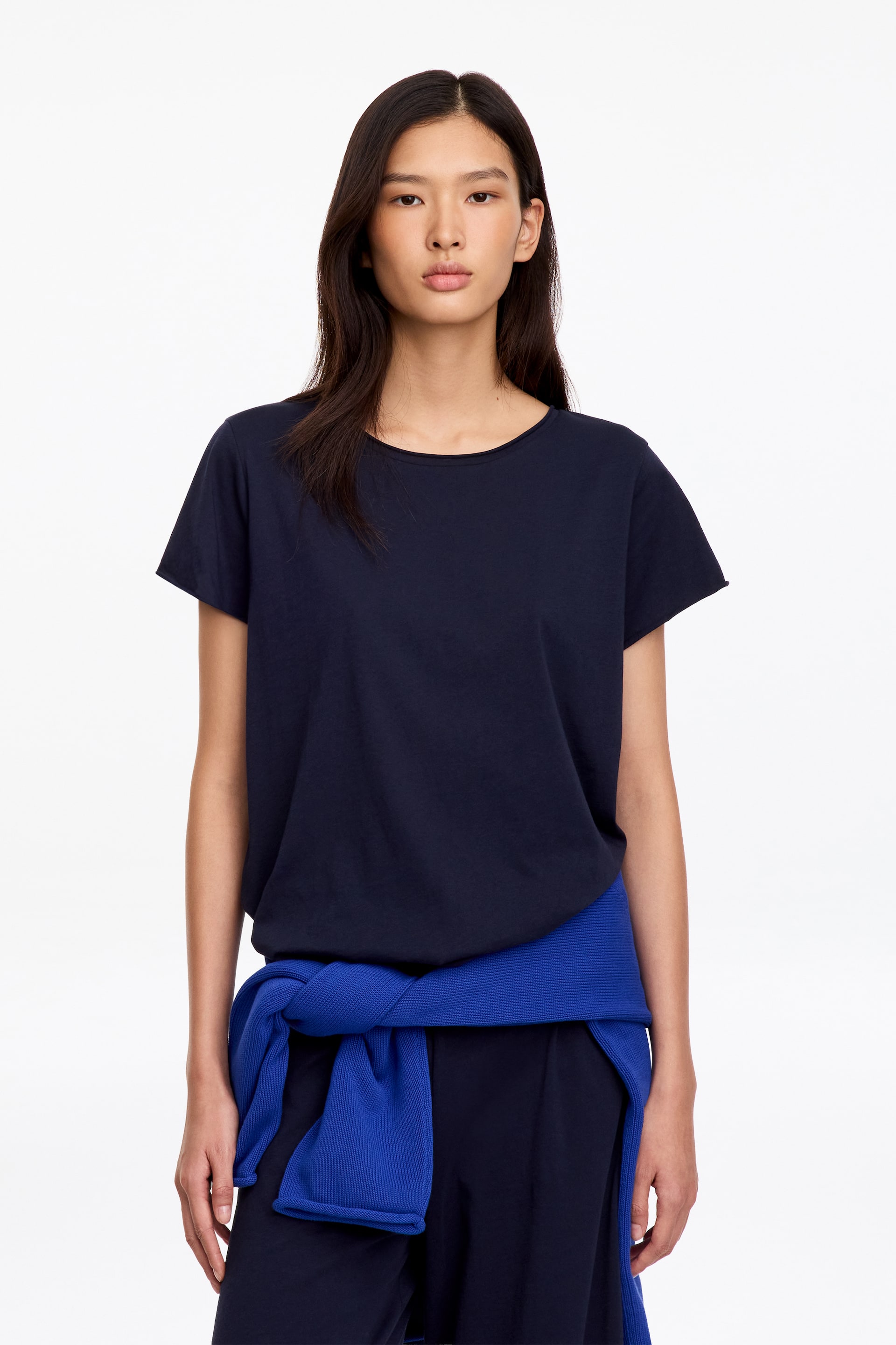 Grösseres Bild ansehen: Ein dunkelmarinefarbenes Jersey-T-Shirt mit lockerer Passform, rundem Ausschnitt und Details mit gerollten Kanten wird vom Model getragen. Das T-Shirt ist mit passenden dunklen Hosen und einem leuchtend blauen Pullover kombiniert, der um die Taille gebunden ist.