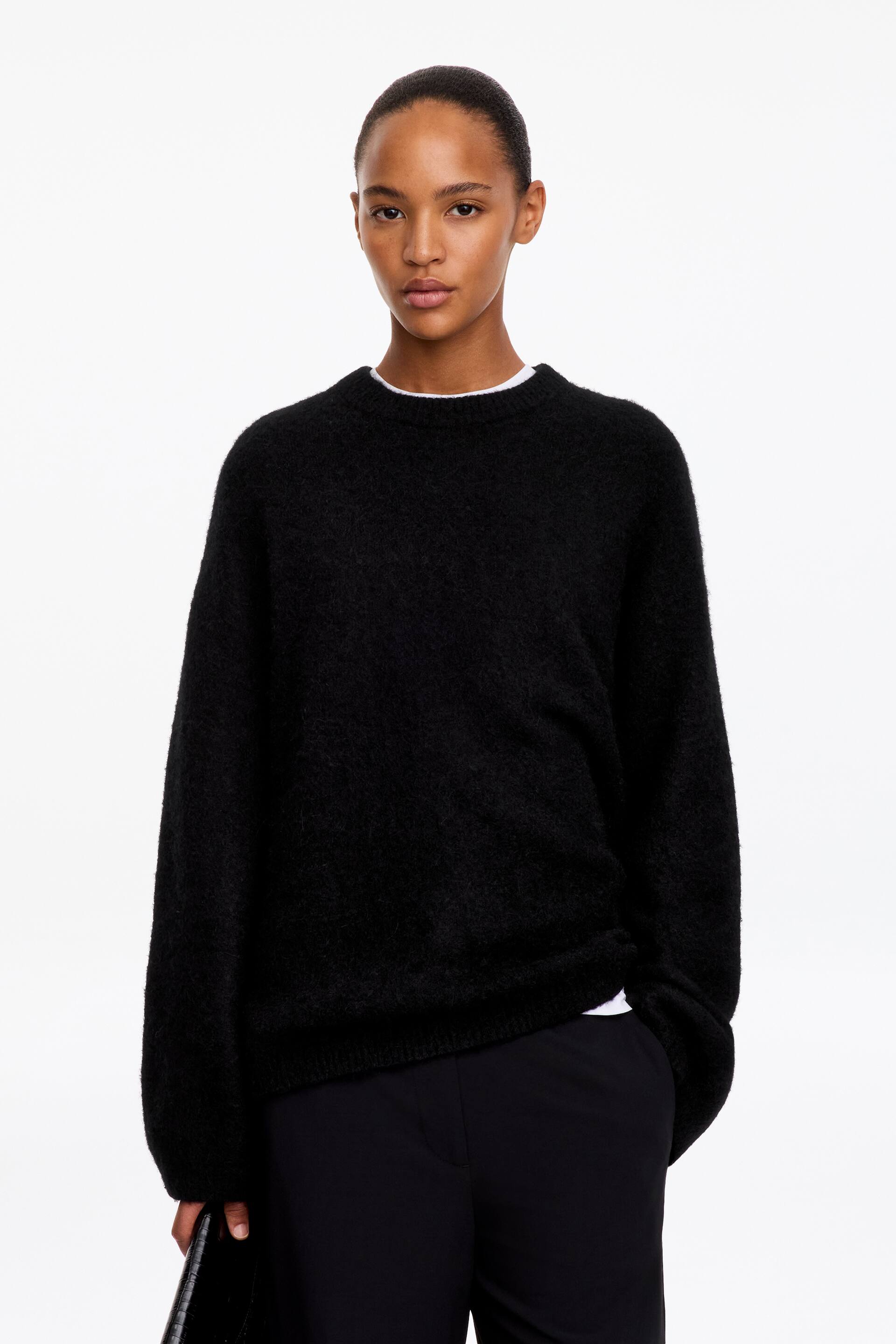 Größeres Bild anzeigen: Ein schwarzer, flauschiger Strickpullover mit Rundhalsausschnitt wird vom Model getragen. Der langärmelige Pullover in lockerer Passform ist über einem weißen T-Shirt getragen und mit einer schwarzen Hose kombiniert.