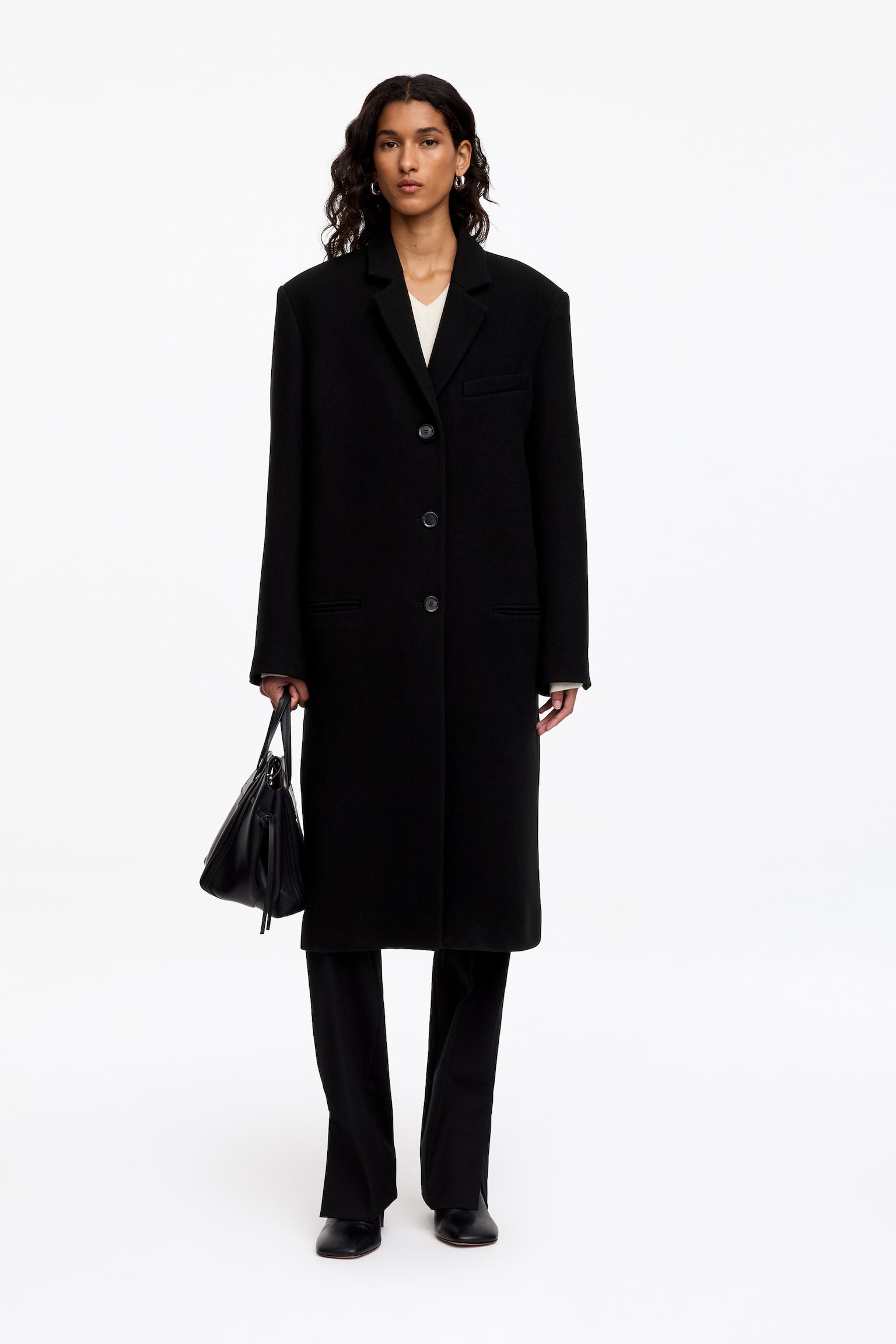 Un manteau noir en mélange de laine, doté dâune coupe à boutonnage simple, de revers crantés et de trois boutons sur le devant, est porté par le mannequin. Ce manteau longueur genou est associé à un haut de couleur claire et à un pantalon noir, le mannequin portant également un sac à main en cuir noir. - 1