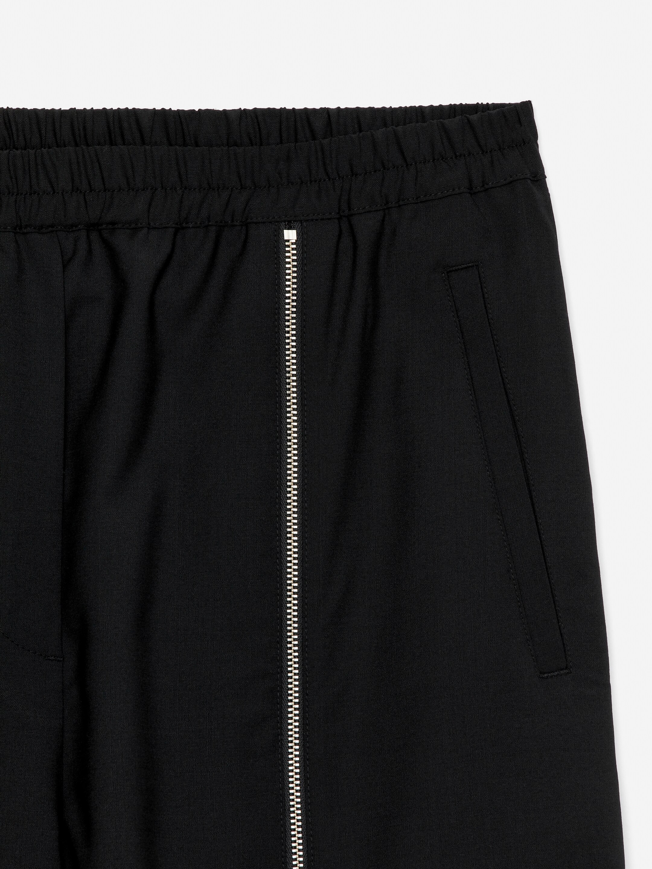 View larger image: Black tailored sweatpants are presented from the front, featuring an elasticated waist. A long silver vertical zipper is running down the leg from the waistband, and a welt side pocket is visible on the wearer's right.
