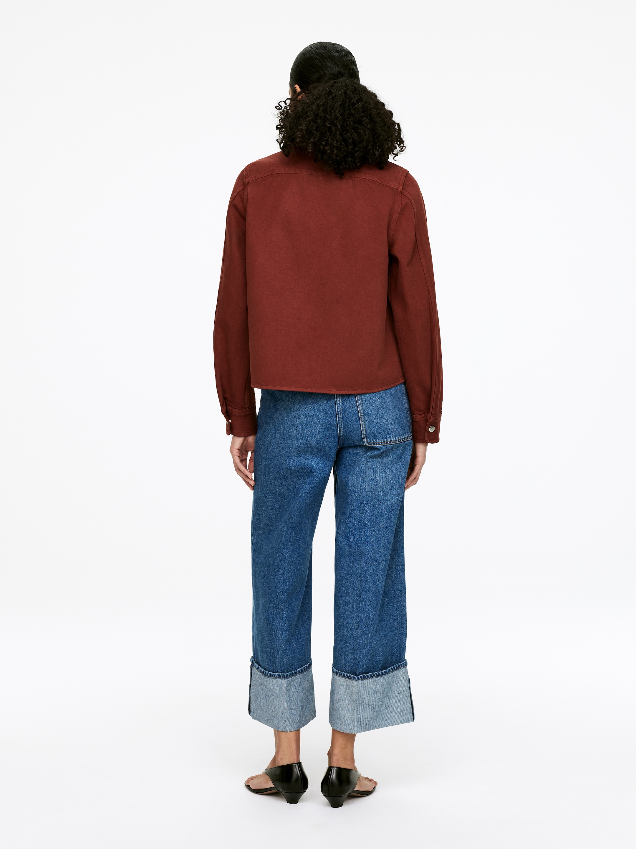 Visualizza immagine più grande: Una overshirt in denim di cotone tinto in capo color marrone-rossiccio è indossata dal modello. Presenta una carcassa posteriore, maniche lunghe e una vestibilità regolare leggermente corta, ed è abbinata a jeans blu in denim con risvolto.
