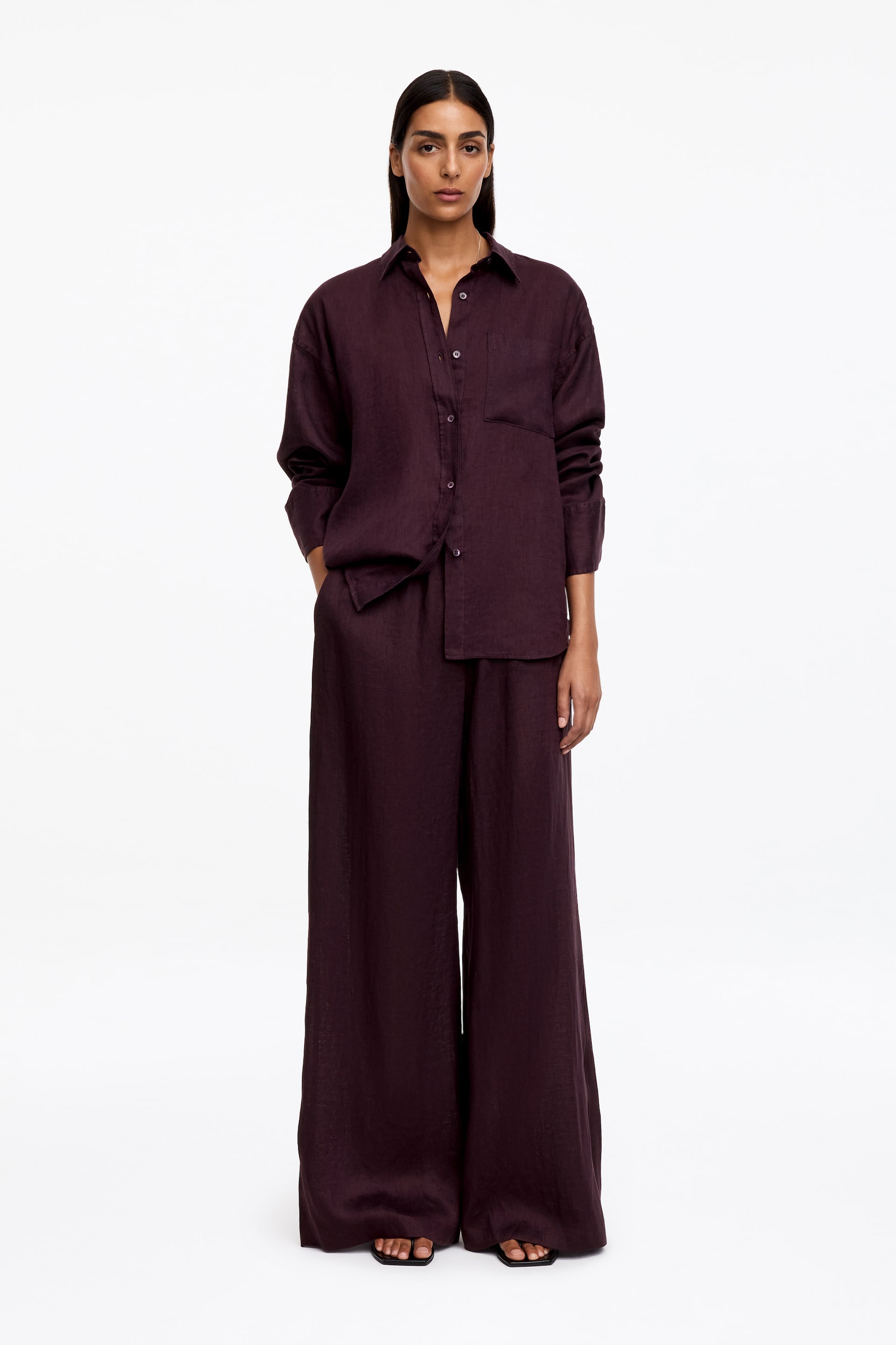 Agrandir l'image: Un pantalon en lin prune foncé, doté de jambes larges et fluides et d’une silhouette décontractée, est porté par le mannequin. Le mannequin porte également une chemise assortie à manches longues, et une main repose dans la poche latérale du pantalon.