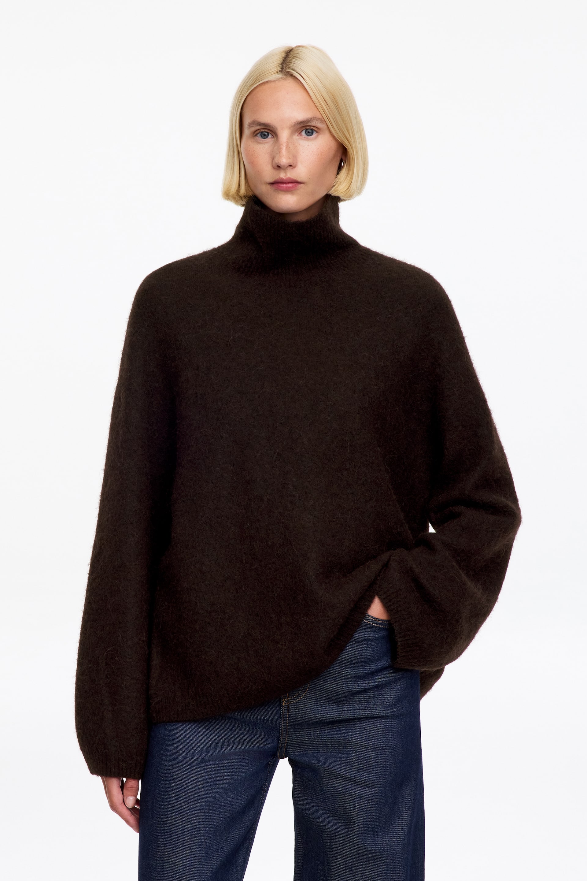 Ver imagen más grande: La modelo lleva un jersey de cuello alto, de color marrón oscuro y corte oversize, con una textura afelpada, mientras mantiene una mano metida en el bolsillo de unos vaqueros azul oscuro. El jersey presenta mangas holgadas con puños de punto acanalado y un cuello alto y suave.