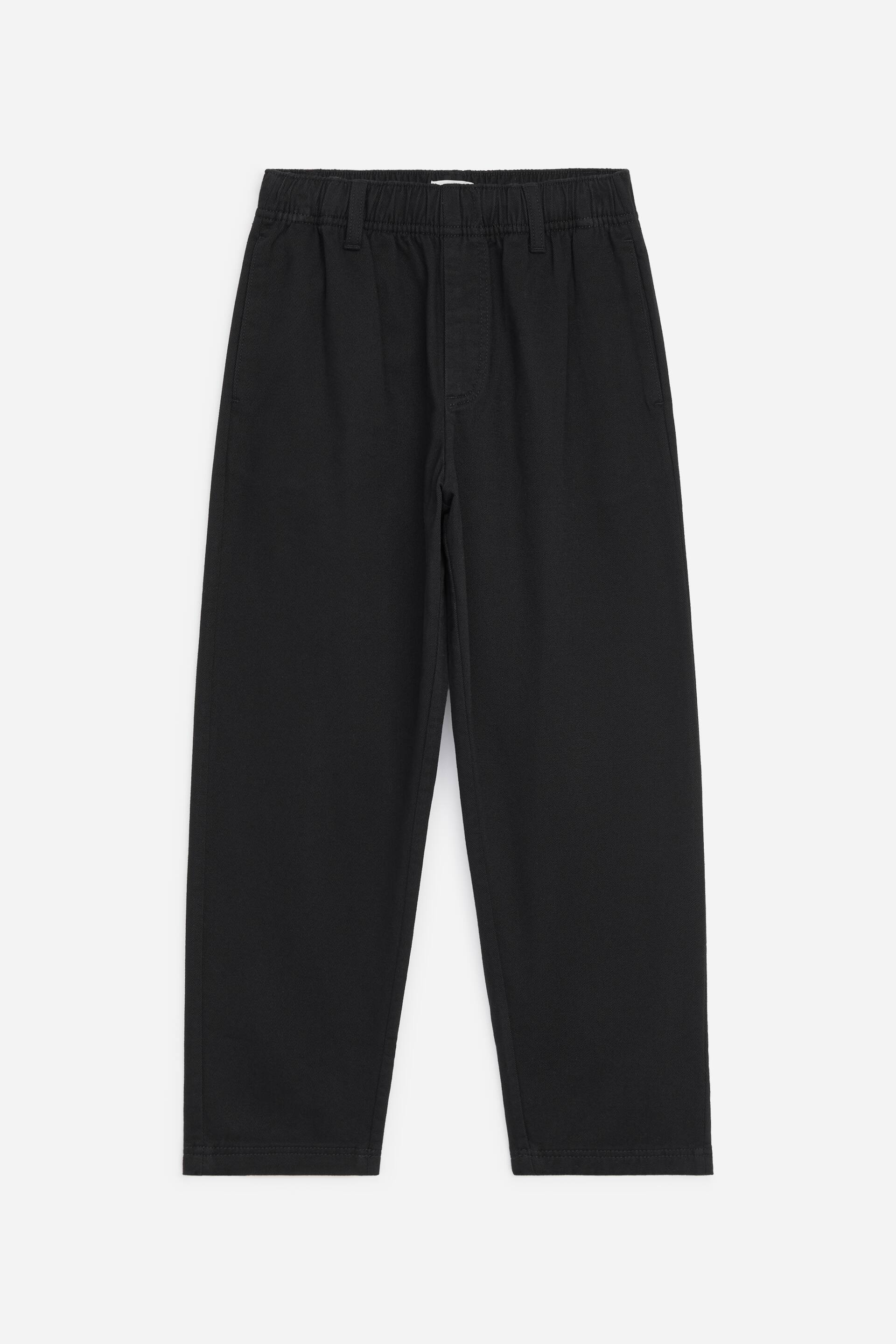 Größeres Bild ansehen: Frontansicht der schwarzen, schmal zulaufenden Baumwoll-Chinos mit elastischem Bund, Gürtelschlaufen und angedeutetem Hosenschlitz. Die Hose zeigt seitliche Eingrifftaschen und eine lässige Silhouette, die zum Saum hin schmaler wird.