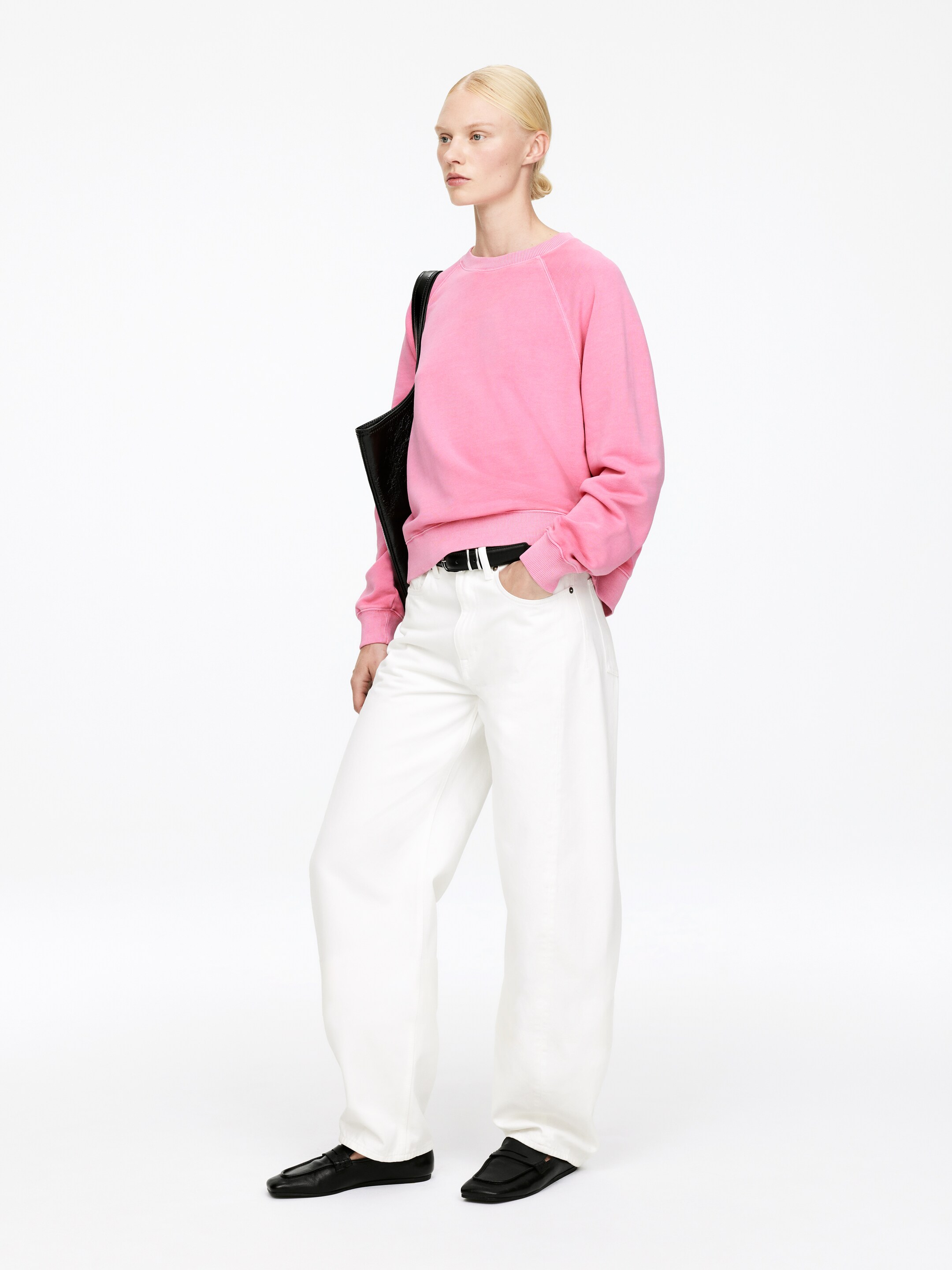 Größeres Bild anzeigen: Ein verblasstes rosa Sweatshirt mit Rundhalsausschnitt, das Raglanärmel, gerippte Bündchen und einen gerippten Saum aufweist, wird mit weißen, weit geschnittenen Hosen getragen, die mit einem schwarzen Gürtel zusammengehalten werden. Schwarze Slip-On-Loafer vervollständigen den Look, während eine schwarze Tragetasche über einer Schulter getragen wird, alles aus einem leicht schrägen Blickwinkel betrachtet.