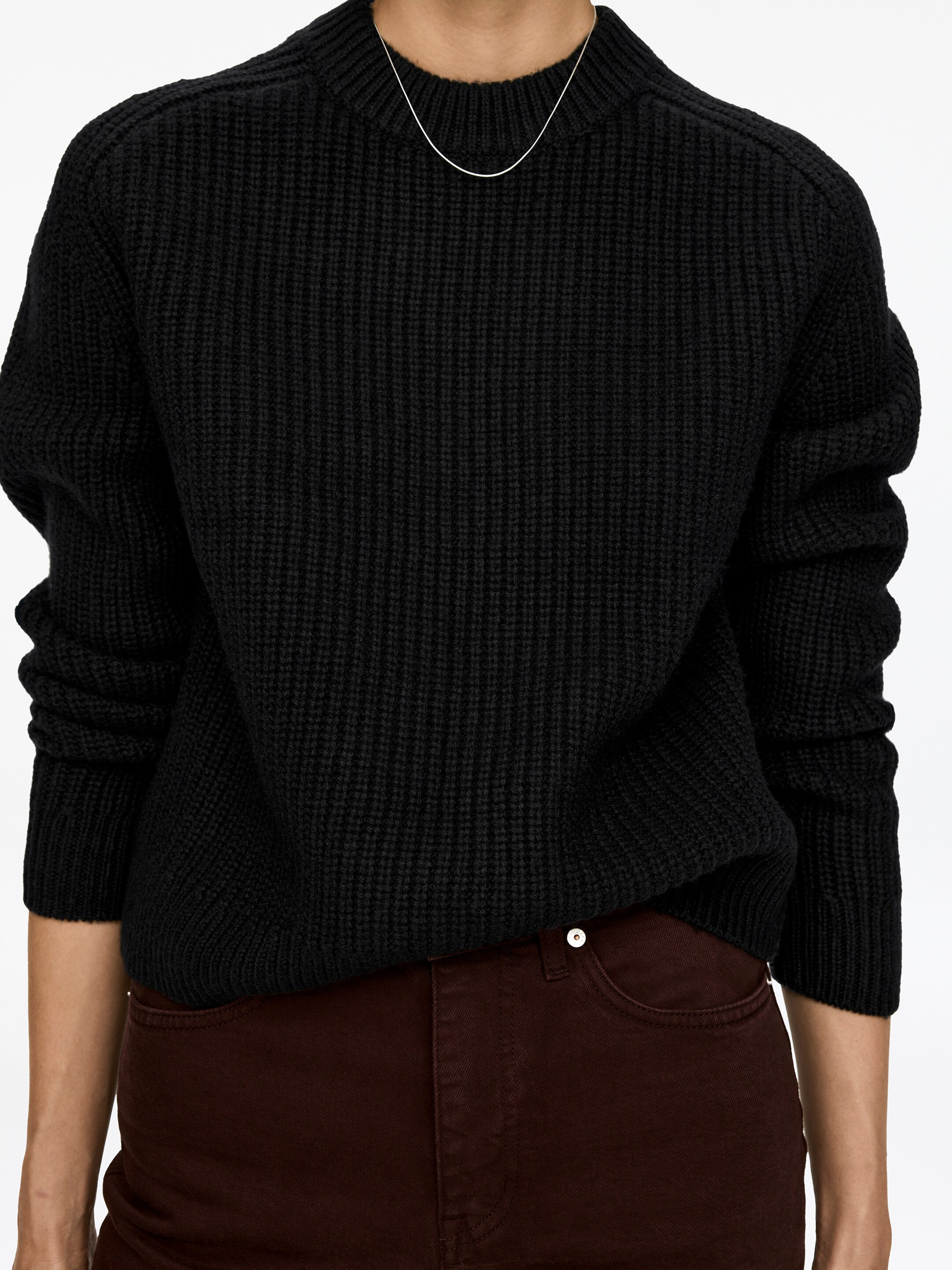 Vis større bilde: En svart genser, strikket i halvpatent med lange ermer, rund hals og ribbede kanter, bæres av modellen. Den er stylet over mørkebrune bukser, og et tynt sølvkjede hviler på genserens rund hals.