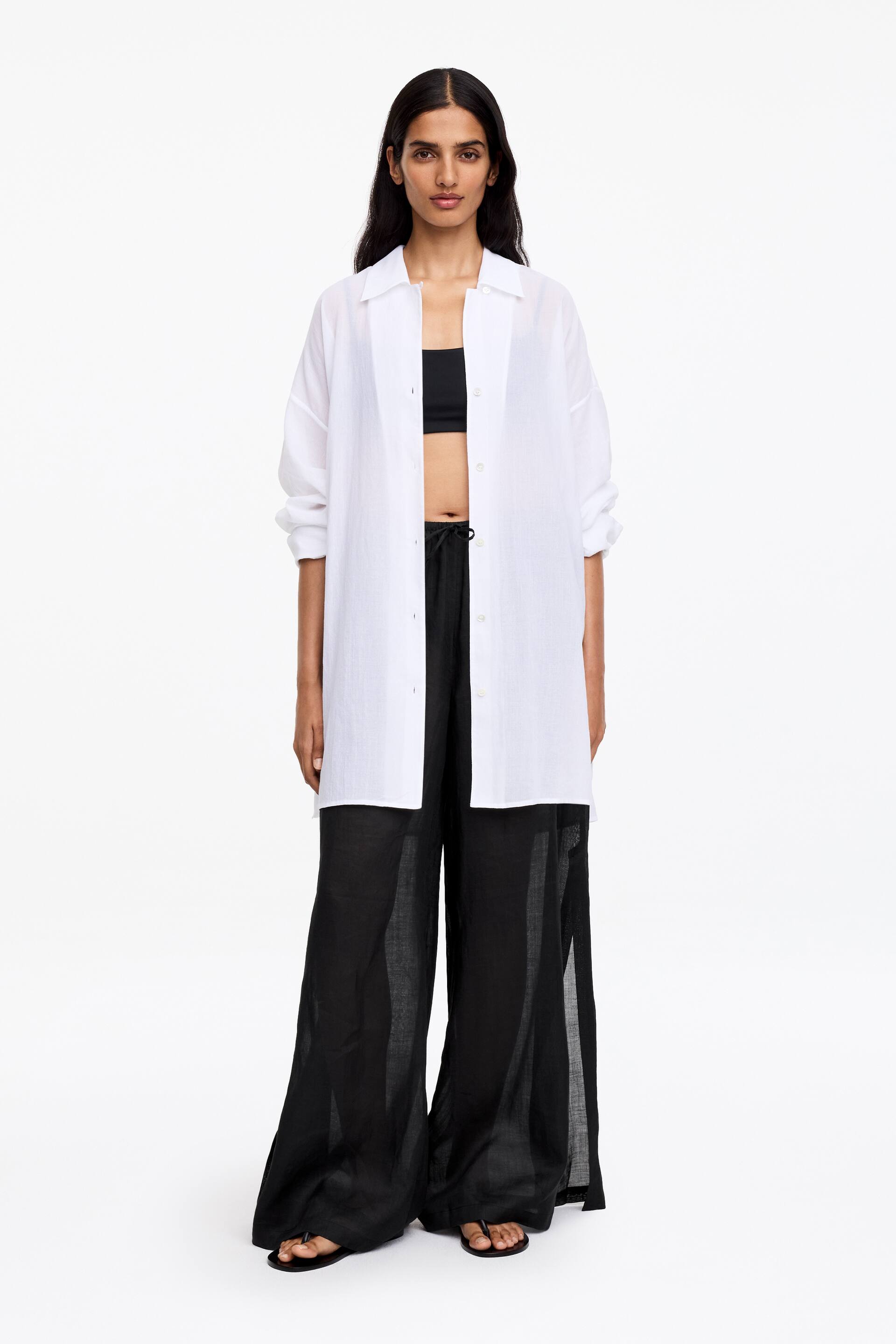 View larger image: An oversized white button-down shirt is styled open over a black bandeau top. It features a classic collar, long sleeves pushed up to the forearms, and a straight hem, and is paired with black wide-leg trousers that display a drawstring waist and a lightweight, sheer texture.
