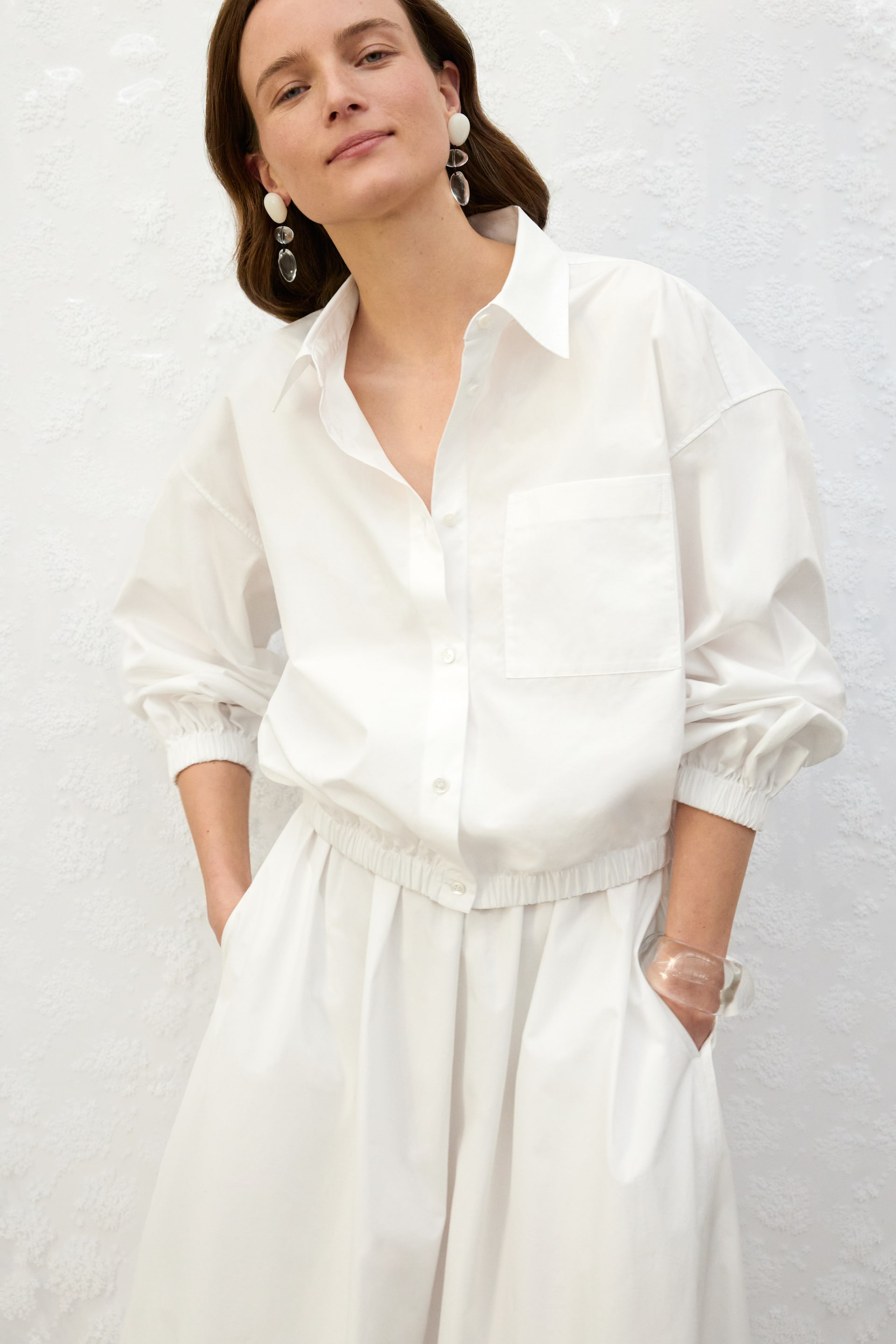 Ver imagen más grande: Una camisa blanca abotonada presenta un cuello en punta, un bolsillo de parche en el pecho y mangas largas con puños elásticos. Su dobladillo elástico descansa justo por encima de pantalones blancos de pierna ancha a juego, que también cuentan con una cintura elástica y bolsillos laterales.