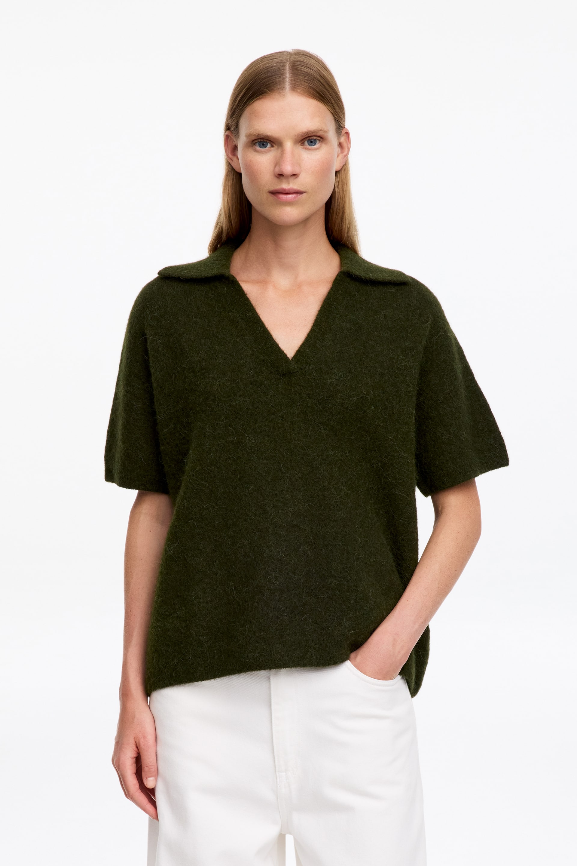 Größeres Bild ansehen: Ein dunkelgrüner, flauschig strukturierter Strick-Polopullover mit lockerer Passform, kurzen Ärmeln und offenem Kragen wird vom Model getragen. Der Pullover ist mit einer weißen Hose kombiniert, wobei das Model eine Hand in einer Tasche hat.
