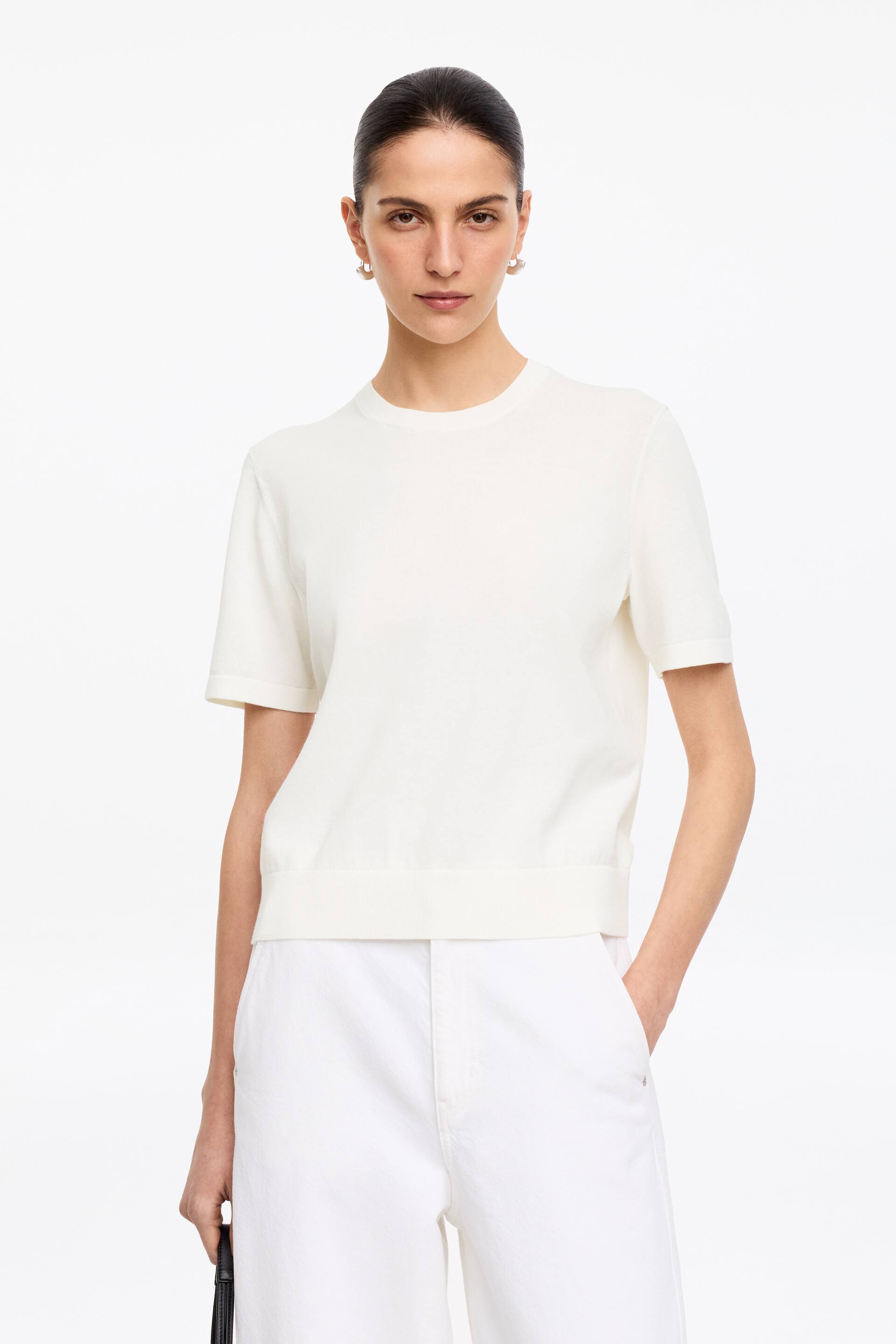 Visualizza immagine più grande: Un top in maglia a maniche corte color bianco sporco è rivolto in avanti, caratterizzato da uno scollo tondo e maniche corte che terminano con polsini a coste sottili. Un'ampia fascia a coste definisce il suo orlo.