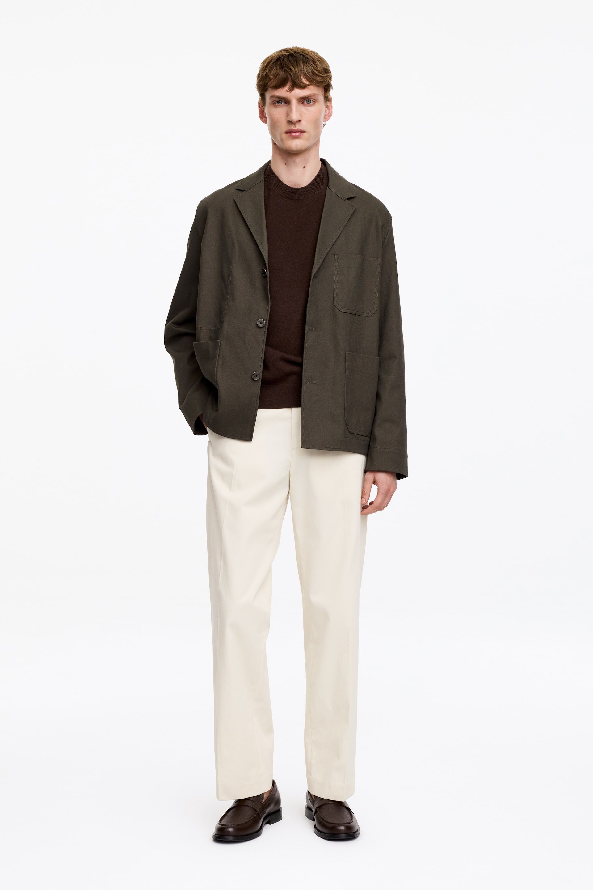 Grotere afbeelding bekijken: Een volledige outfit wordt gepresenteerd, met een donker olijfgroene ongevoerde blazer met inkeping revers en drie opgestikte zakken, gedragen over een donkerbruine trui met ronde hals. De outfit wordt compleet gemaakt met licht crèmekleurige rechte broek en donkerbruine loafers.