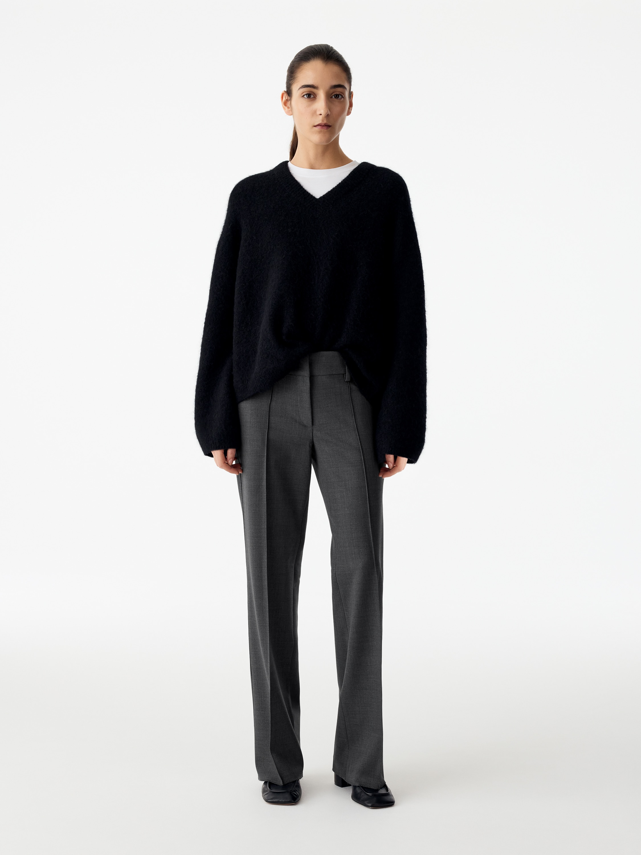 A pair of dark grey tailored trousers is shown from the front, featuring a low-rise waist with belt loops and a concealed fly fastening. The trousers are designed with a slim fit and prominent sewn creases running down the centre of each slightly flared leg.