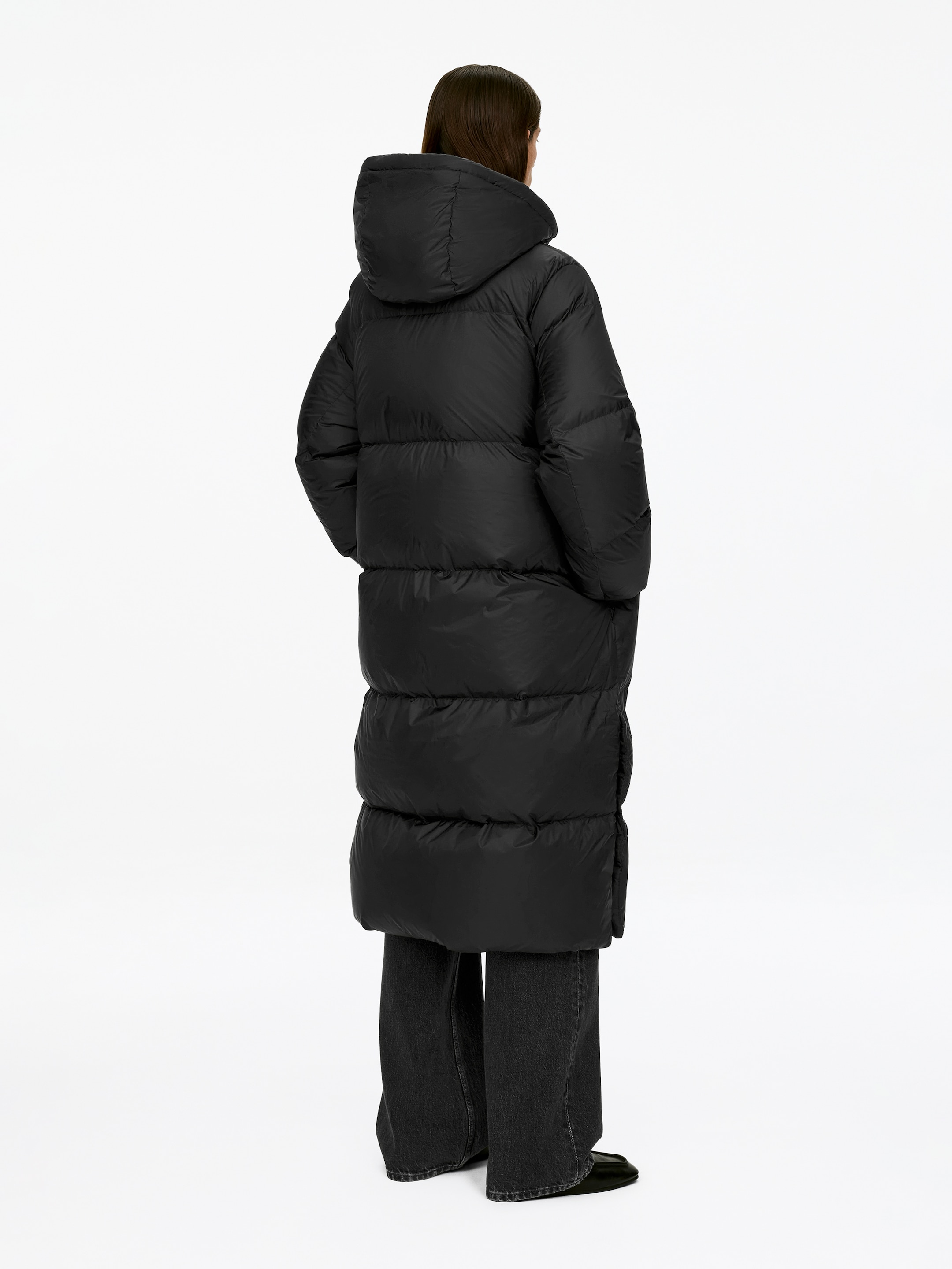 View larger image: The Upcycled Down Long Coat in black is being worn by the model, who is standing with their hands in its pockets. The hooded puffer coat is displaying horizontal quilting and is extending to a long length, with dark trousers being visible beneath its hem.