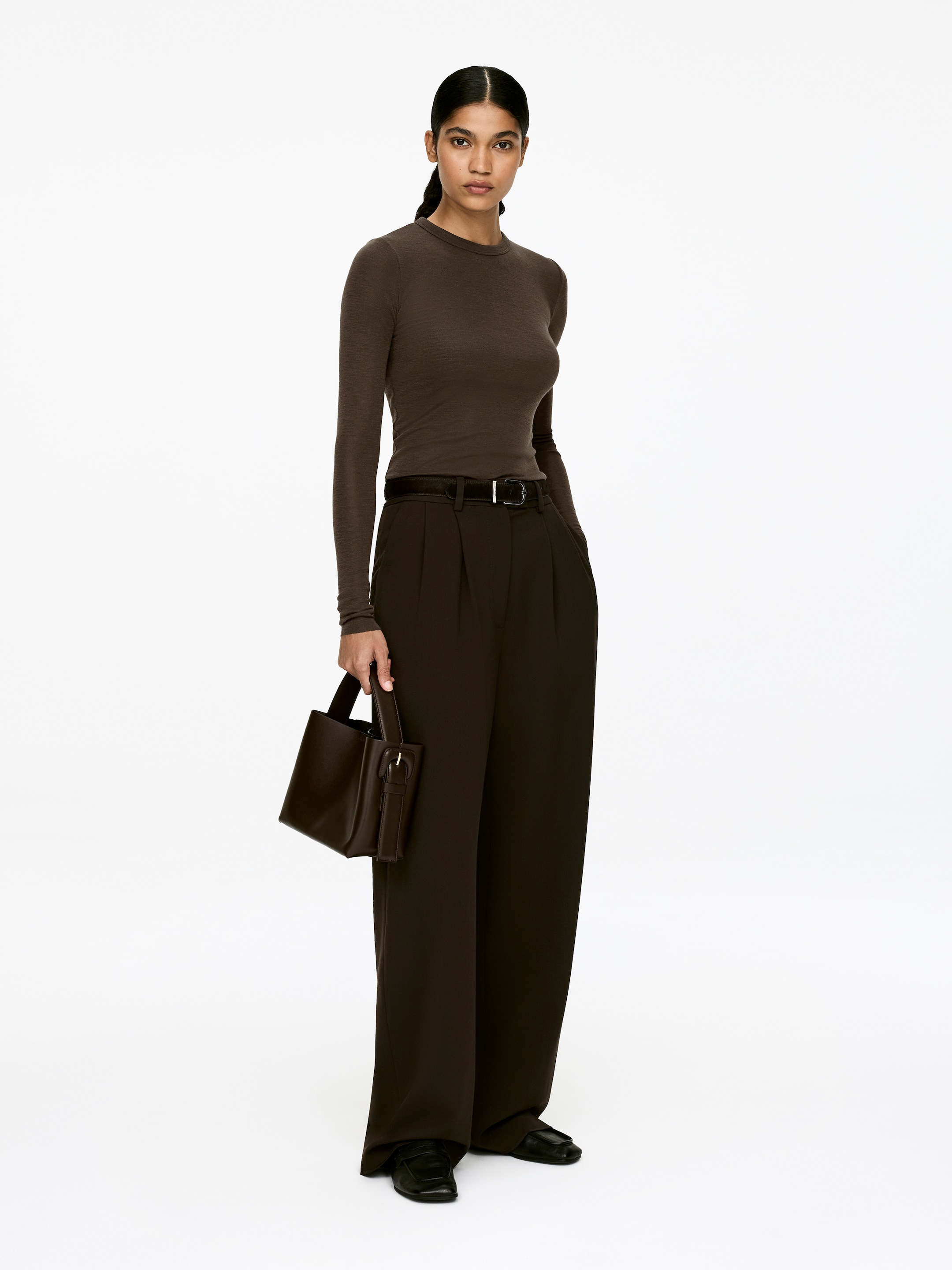 View larger image: A dark brown, tight-fitted merino wool top with a slightly transparent appearance, crew neck, and long sleeves is being worn by the model. The top, featuring raw-cut edges at the hemline and sleeve ends, is tucked into dark trousers.