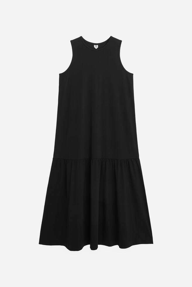 Ein schwarzes ärmelloses Kleid mit einem Rundhalsausschnitt und einem kleinen weißen V-förmigen Emblem auf der Vorderseite. Das Kleid ist mit einer lockeren Passform im Oberteil gestaltet und geht in einen gerafften Volant am Saum über. - 1