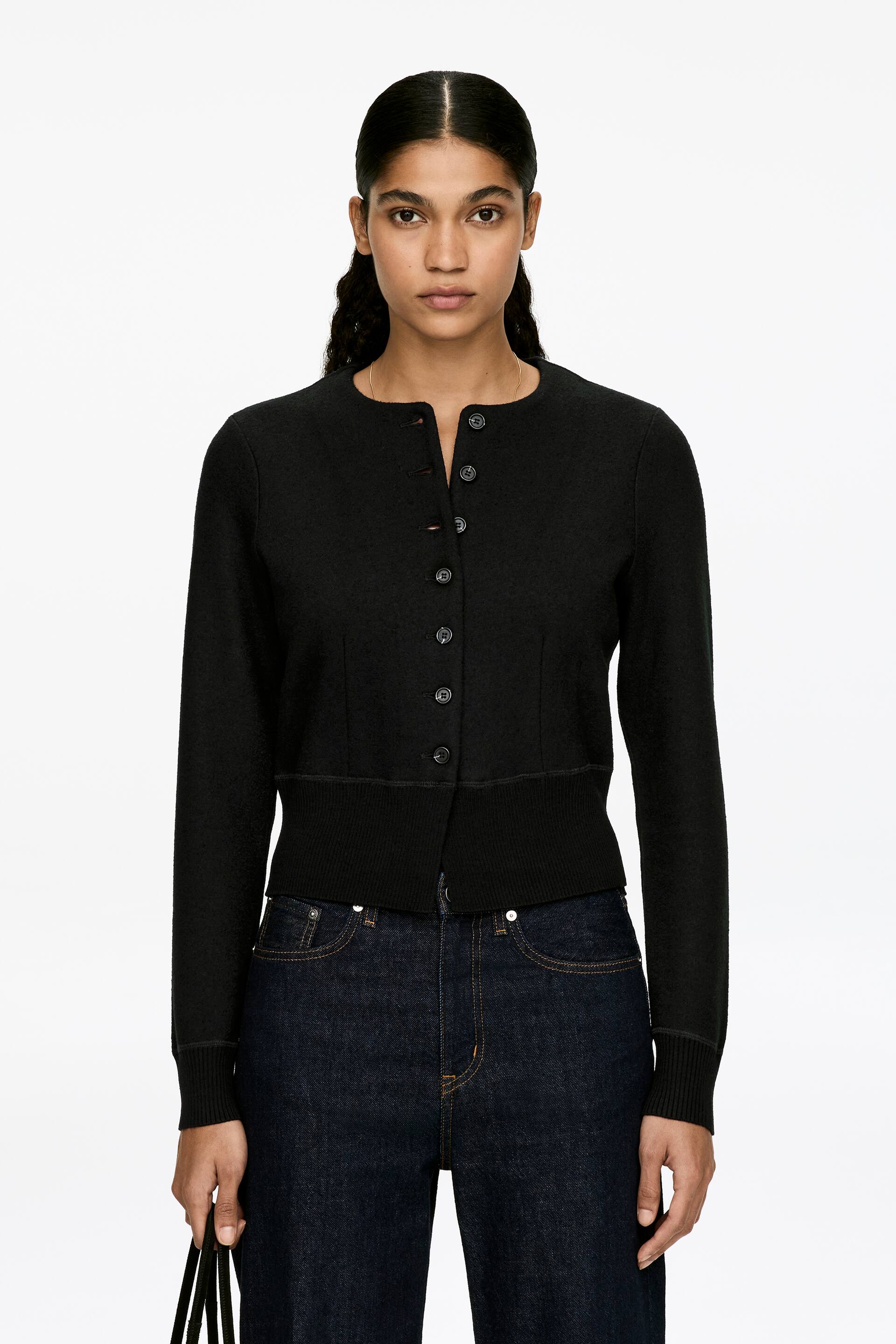 Vis større bilde: En svart cardigan med en knappeåpen front, med mørke knapper og noen åpne øverste knapper, bæres av modellen. Cardiganen viser en slank passform med synlige midjebrett som skaper en tettsittende silhuett. Avsluttende detaljer på den ribbestrikkede kanten og mansjettene, og modellen holder også en svart veske.