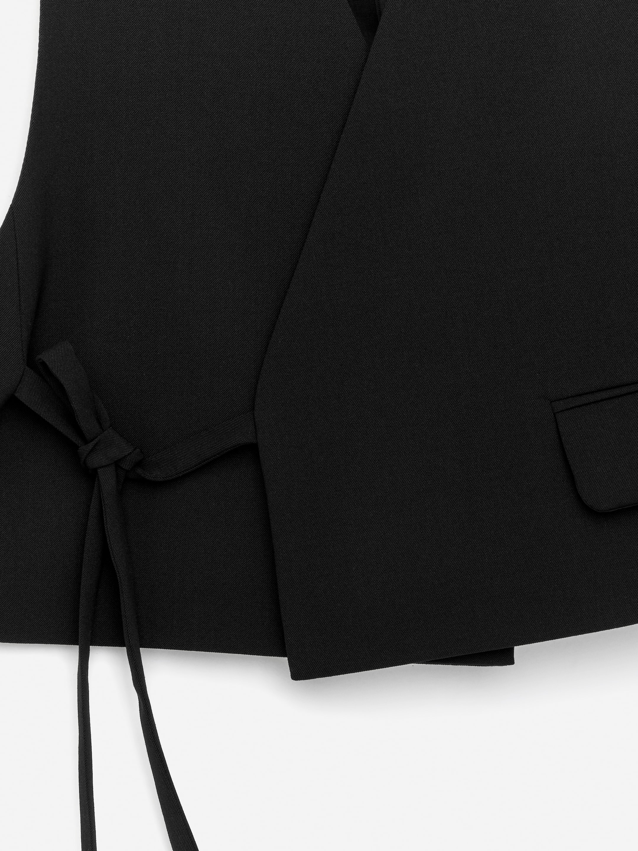View larger image: A black tailored vest is shown from the front, featuring an overlapping design. It is being secured by a tie strap that is knotted on the left side, and a pocket flap is visible on the right.