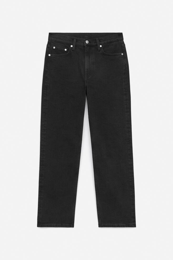 Een paar zwarte denim jeans wordt plat gepresenteerd, met de voorkant naar voren. Ze hebben een middelhoge taille, een zichtbare ritssluiting en twee voorzakken met metalen klinknagels. De pijpen lijken slank, taps toelopend en kort. - 1