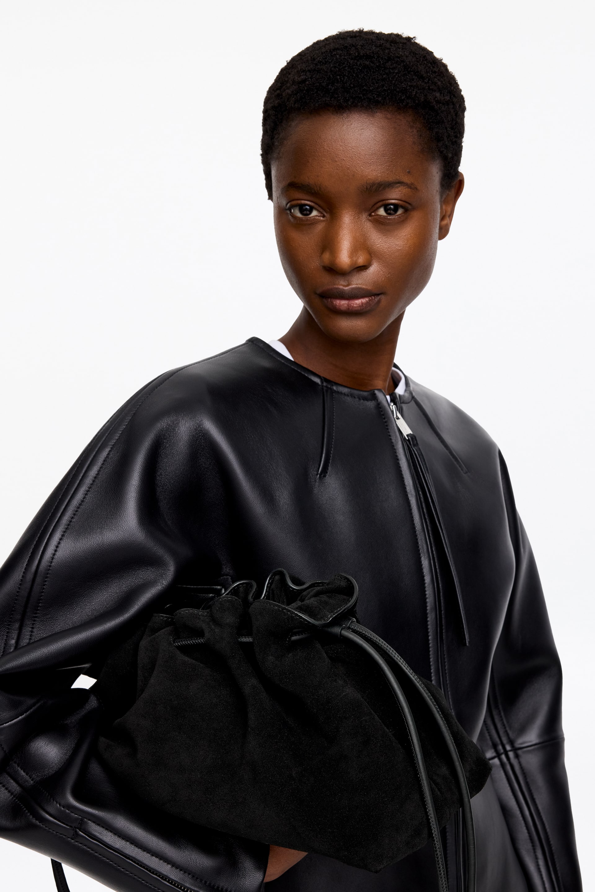 Grösseres Bild ansehen: Eine schwarze Lederjacke mit kragenlosem, rundem Ausschnitt und einem Frontreißverschluss, der hoch auf der Brust endet, mit markanten Nahtlinien an den Ärmeln. Eine schwarze Wildleder-Tasche mit Kordelzug wird vorne gehalten und zeigt ihren gerafften oberen Teil und die dünnen Kordelzüge.