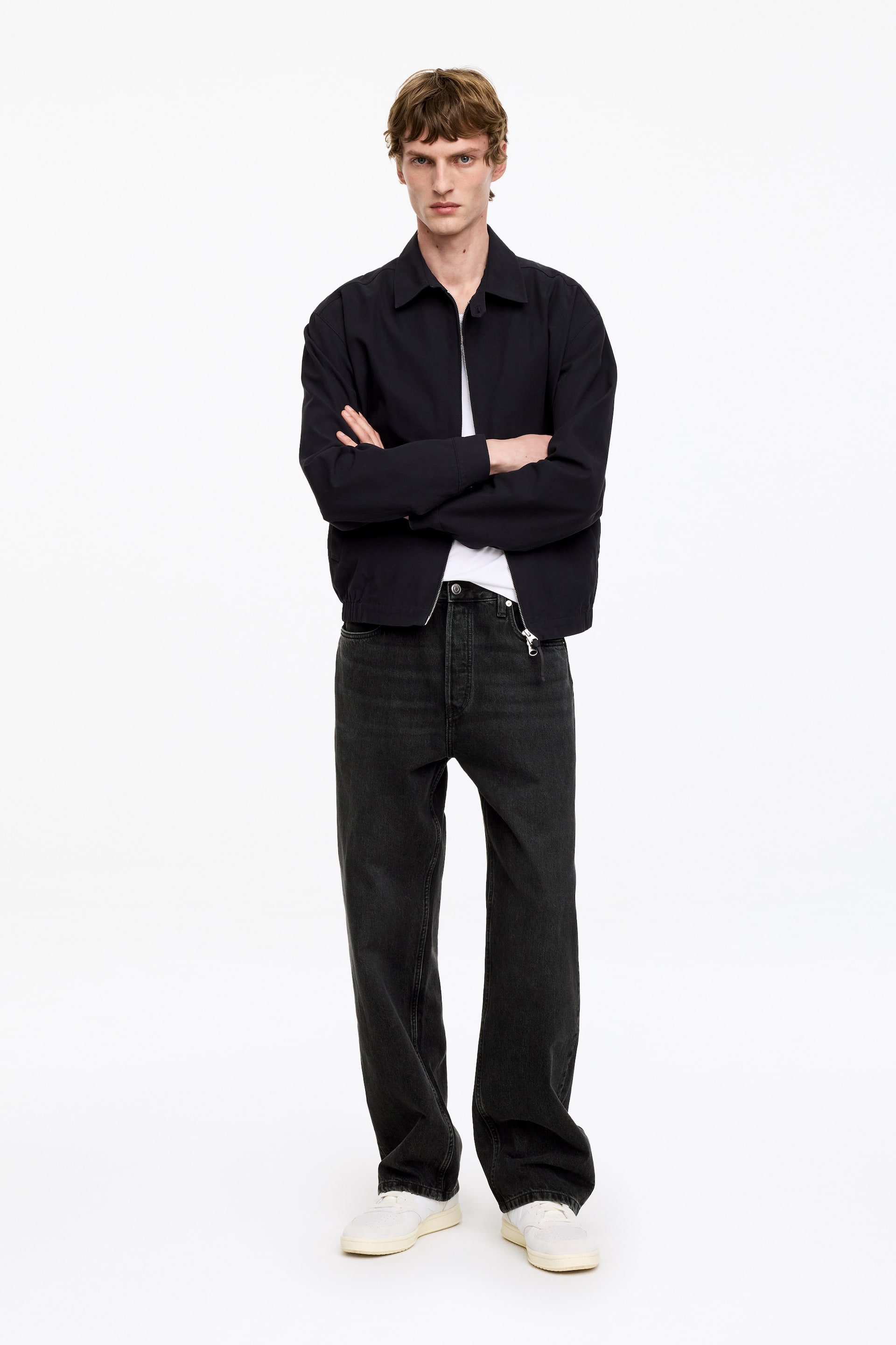 Se større billede: En mørk marineblå jakke med krave bæres, som har en synlig frontlynlås og en elastisk kant. Denne jakke er lagdelt over en hvid crewneck t-shirt, og mørkegrå jeans med afslappet pasform kombineres med hvide lave sneakers.