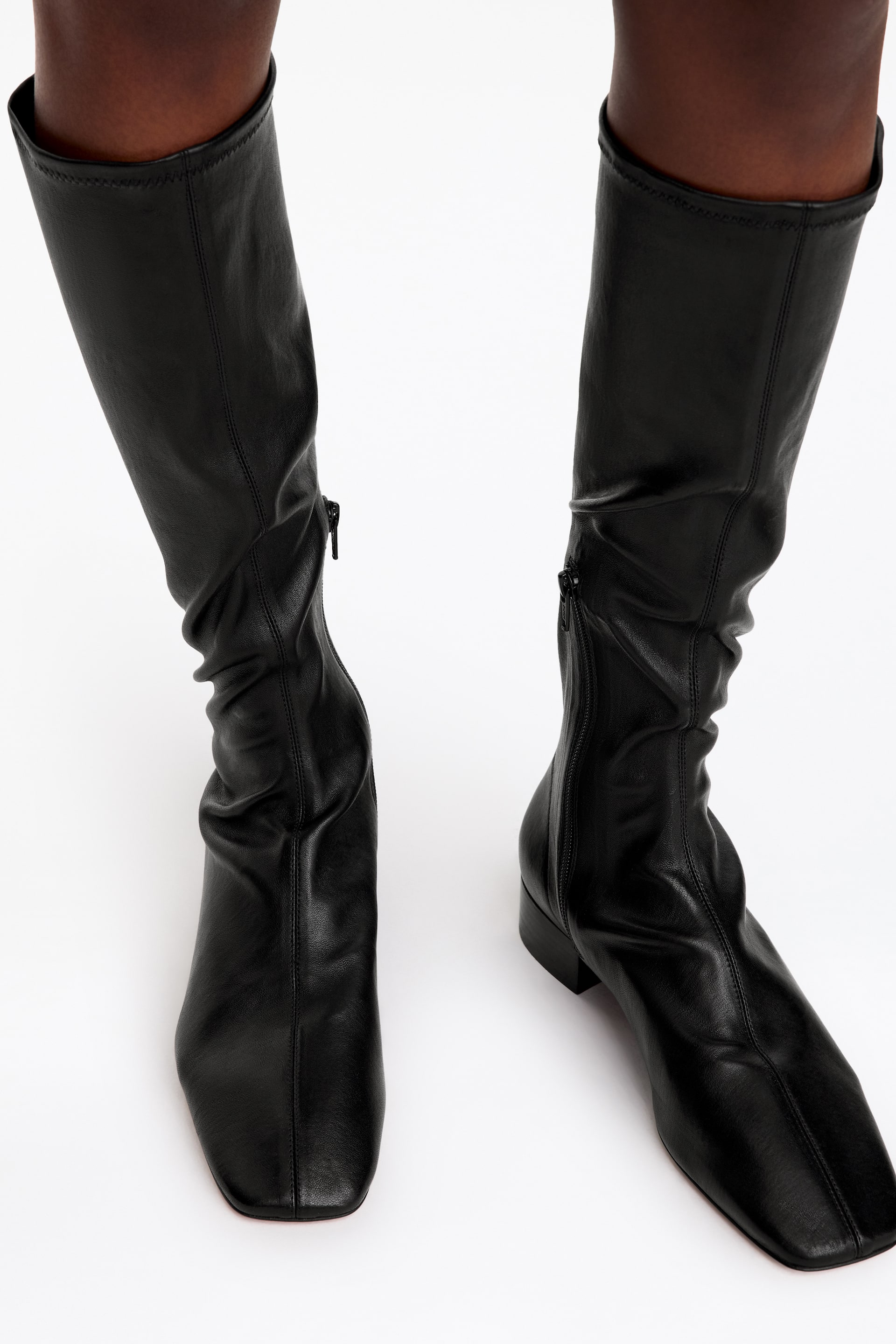 View larger image: Black leather below-the-knee boots with a close-fitting stretch construction are being worn by the model. The boots feature square toes, a central seam along the front, low block heels, and half-zips on the inner sides.