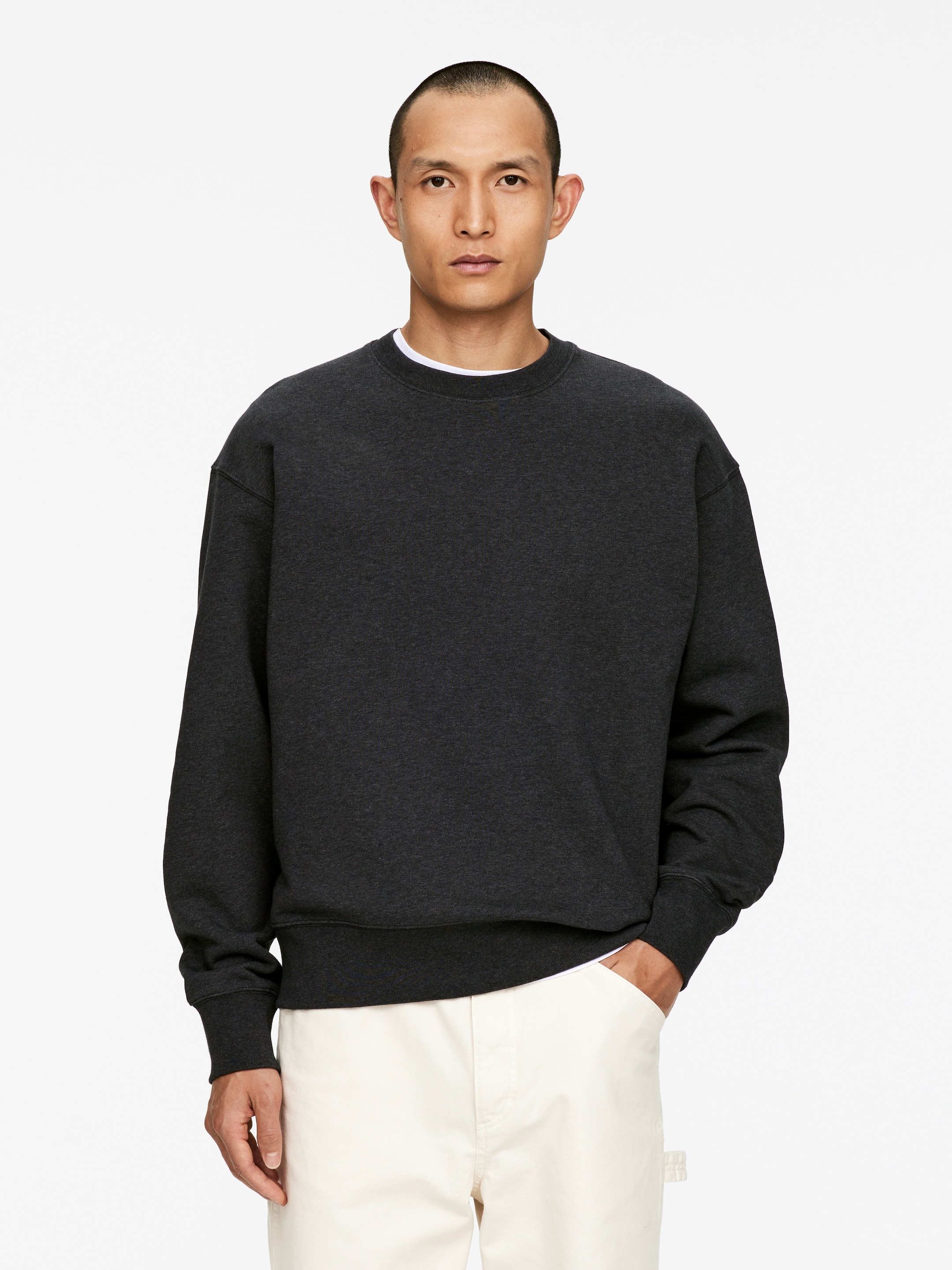 Größeres Bild ansehen: Ein dunkelgraues, meliertes Sweatshirt mit Rundhalsausschnitt wird getragen, nach vorne gerichtet, mit langen Ärmeln mit gerippten Bündchen und einem gerippten Saum. Ein weißes T-Shirt ist unter dem Rundhals sichtbar.