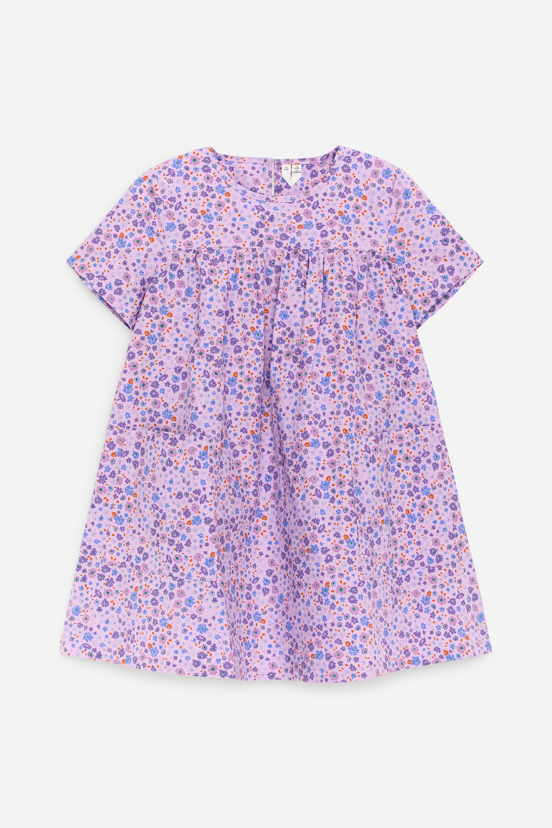 Visa större bild: En lila sommarklänning visas i FRONTAL vy, med ett blommigt tryck i lila, blå och orange nyanser. Den är designad med korta ärmar, en rund halsringning, ett ok upptill med veck under som skapar en voluminös siluett, och två påsydda fickor framtill.