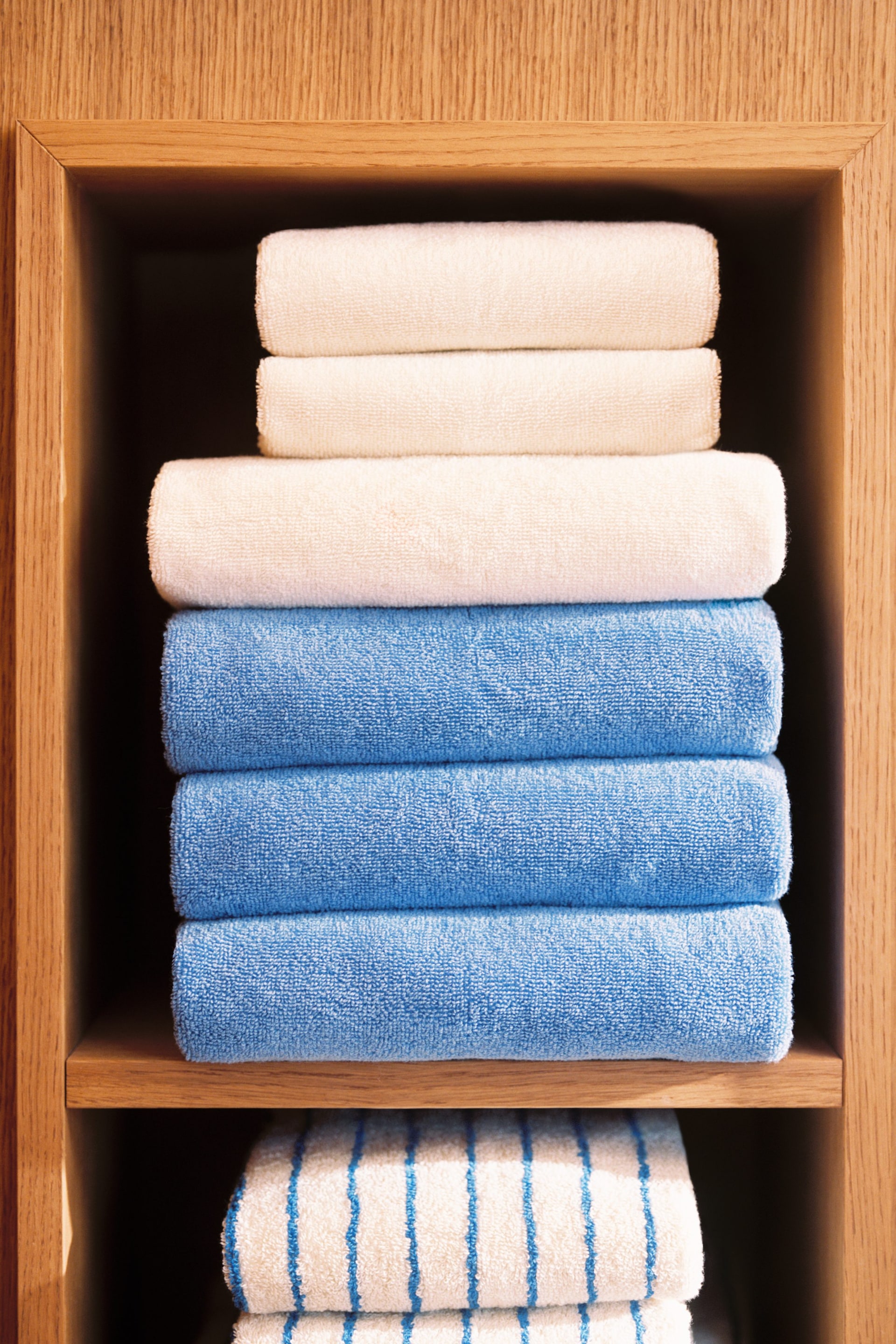 View larger image: Terry cloth towels are displayed, neatly folded and stacked, with their folded edges facing forward. The stack features three cream-coloured towels at the top, three solid blue towels in the middle, and blue and white striped towels partially visible at the bottom.