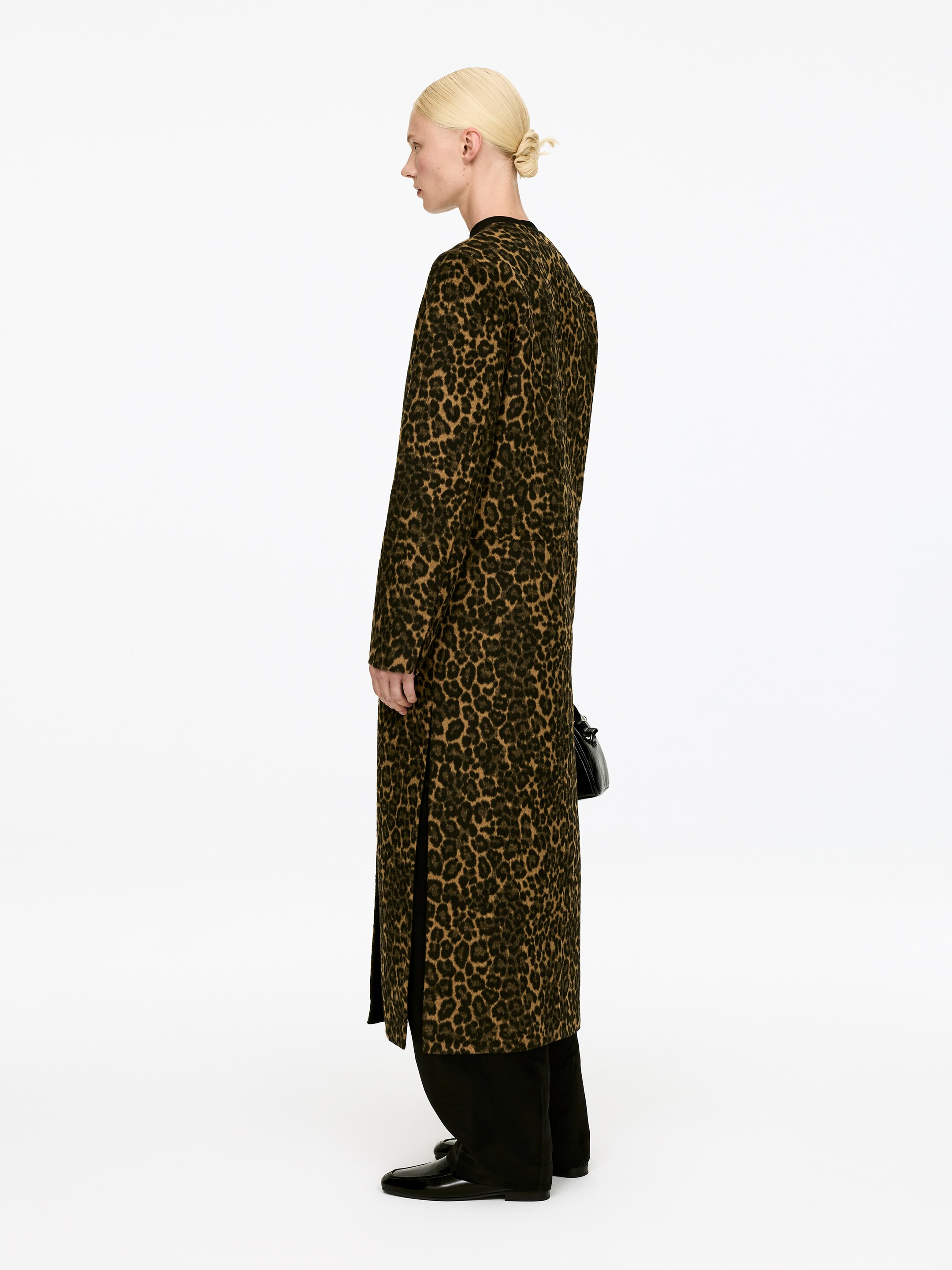 Vis større bilde: En lang frakk med et brunt og svart leopardmønster vises fra siden med ansiktet mot høyre. Den er designet med lange ermer og en rett, ankel-lang silhuett, og har sidespalter som avslører svarte bukser under.
