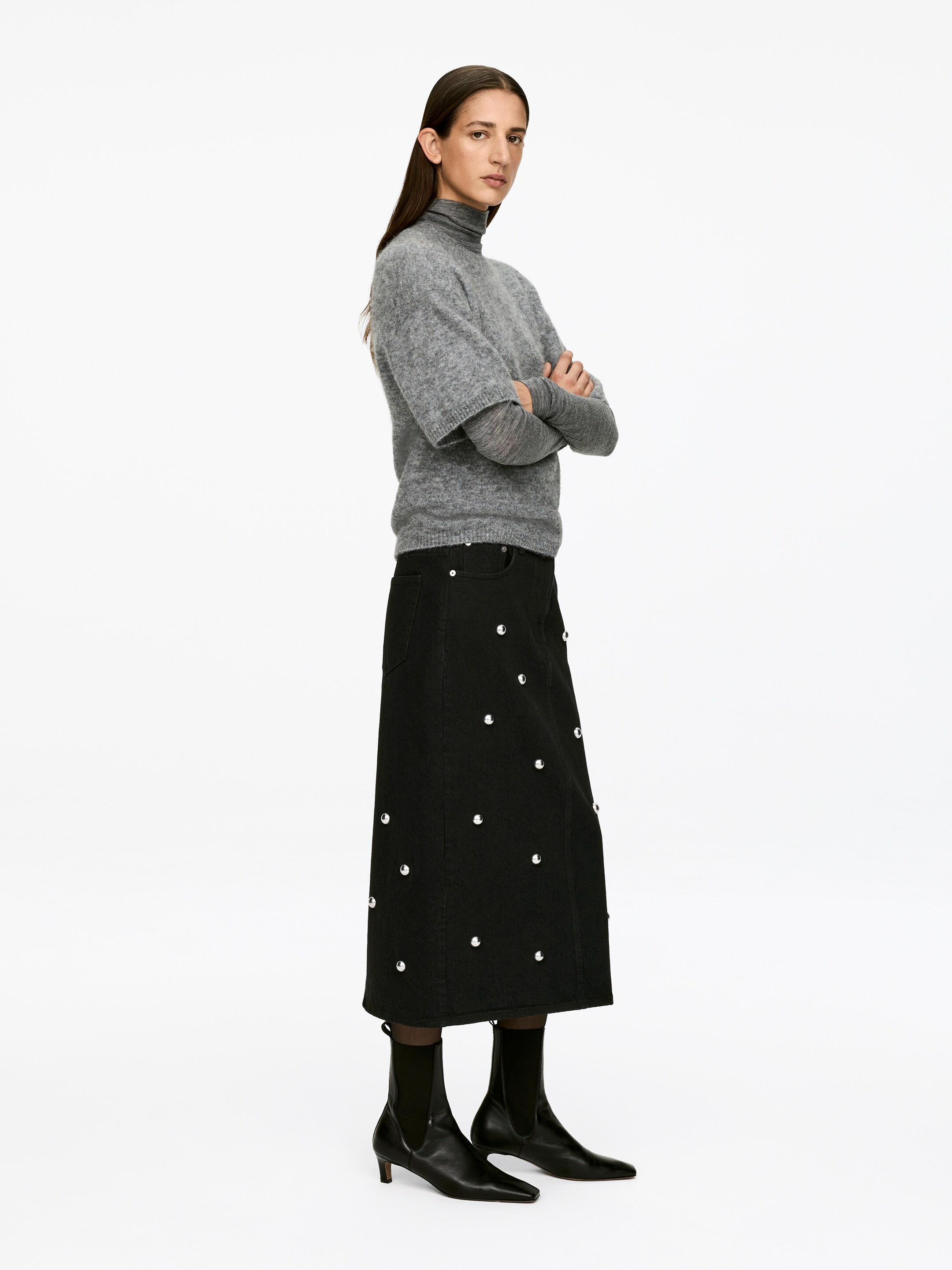 Agrandir l'image: Une jupe mi-longue en coton noir, avec un design à cinq poches et des clous argentés, est portée par le mannequin. La jupe, présentant une coupe régulière, est assortie d'un pull à col roulé gris et de bottes noires.