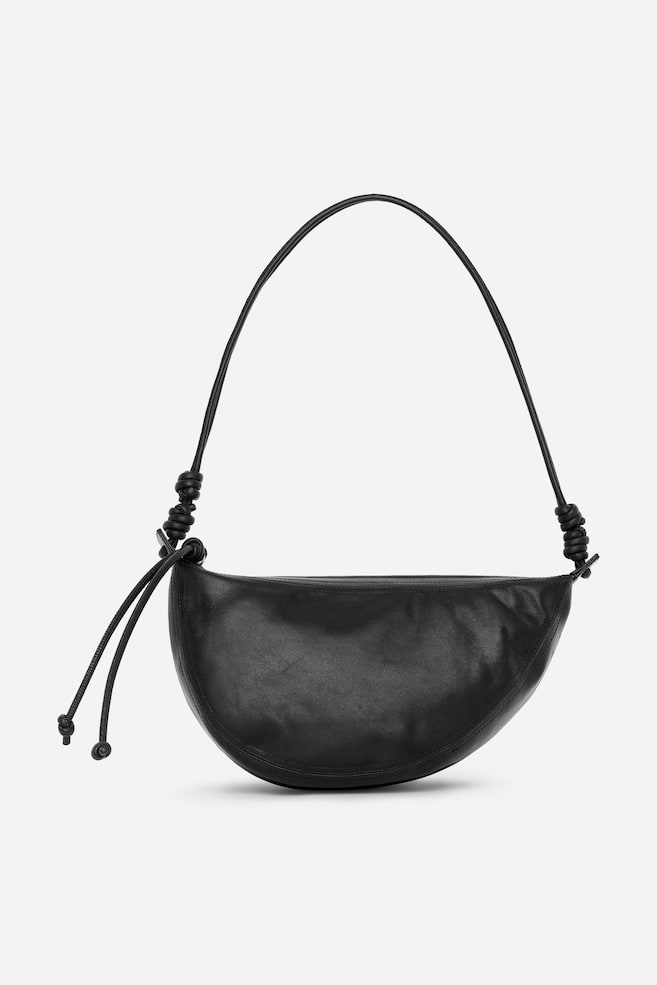 Un sac à bandoulière en cuir noir en forme de croissant est orienté vers l'avant. Il est doté d'une fine sangle en forme de cordon avec des attaches nouées de chaque côté. Une fermeture éclair est visible le long du bord supérieur du sac, et deux tirettes cordées avec des extrémités sphériques pendent du côté gauche. - 1