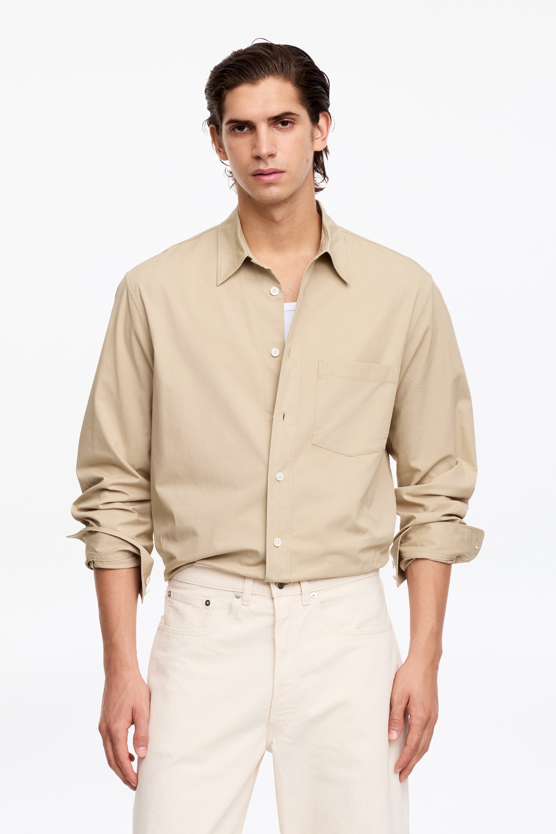 Grösseres Bild ansehen: Das Model trägt ein beige Langarmhemd mit klassischem Kragen, Knopfleiste vorne und einer Brusttasche; die Ärmel sind hochgekrempelt. Das Hemd ist über einem weißen Unterhemd getragen und mit hellen Hosen kombiniert.